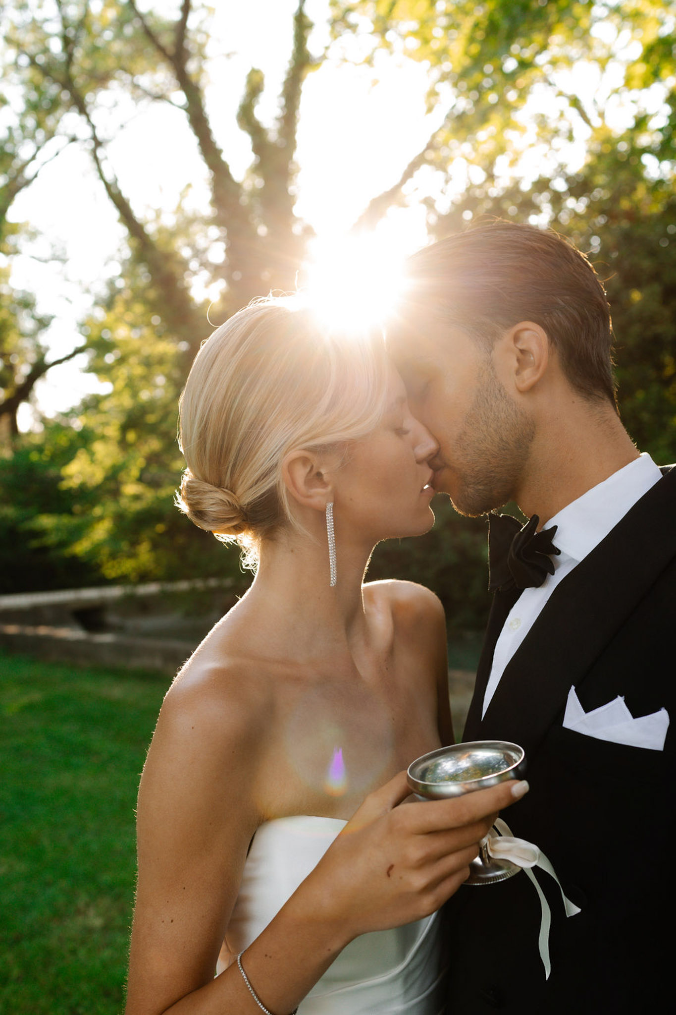 Bride and groom sharing a kiss at outdoor garden reception in profile against backlit trees, golden hour light