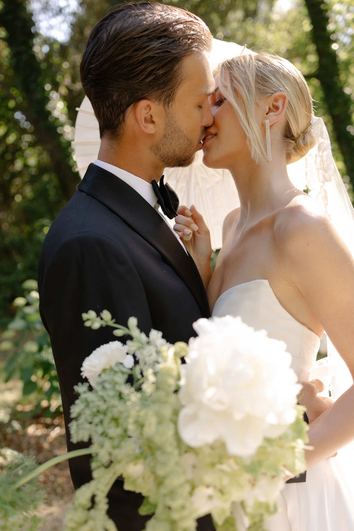 Bride and groom sharing a kiss in garden setting, bride in strapless ivory gown with veil and white bouquet