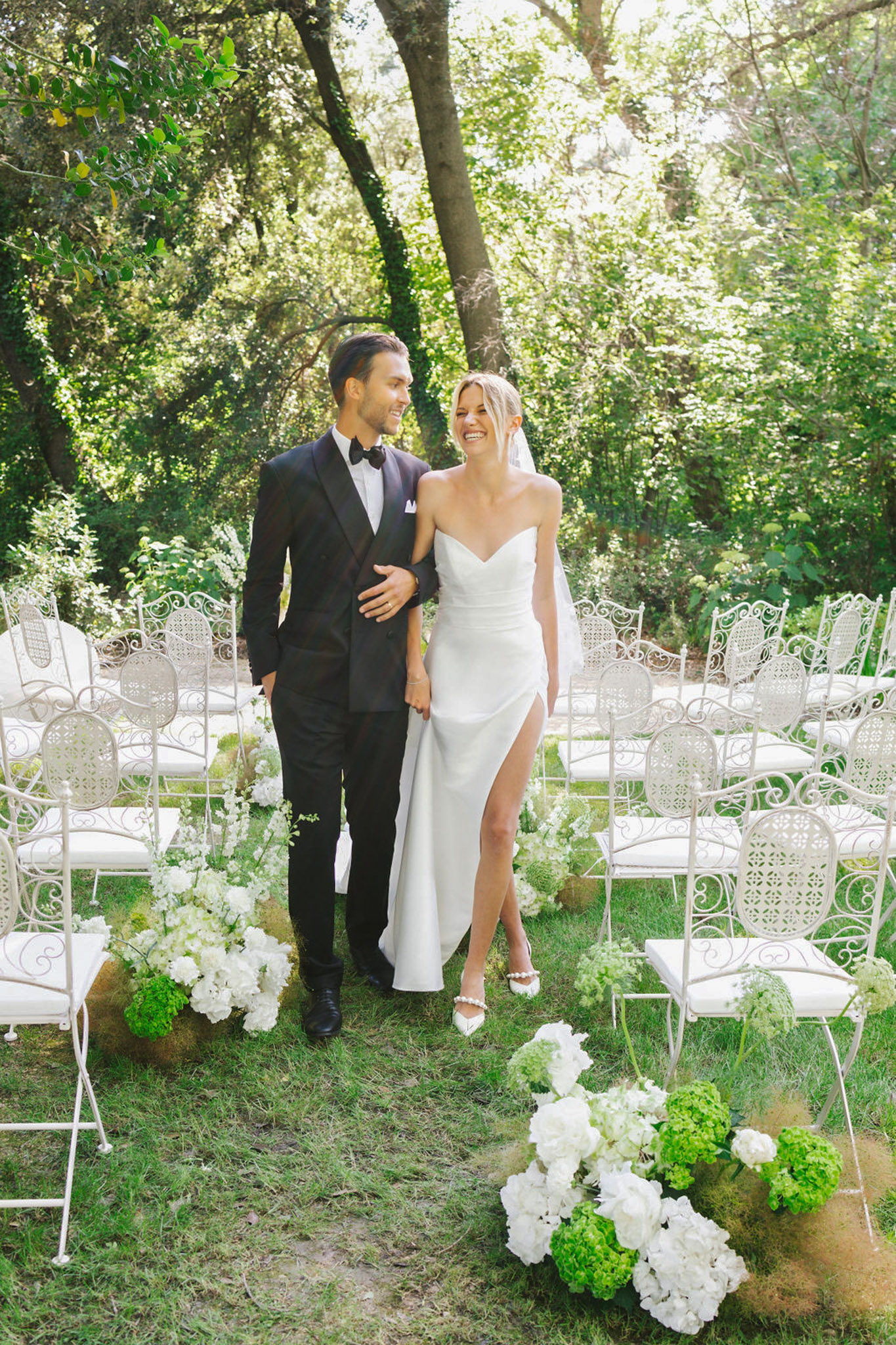 Bride in sleeveless ivory dress with veil and groom in black tuxedo walk aisle lined with white hydrangeas under tree canopy