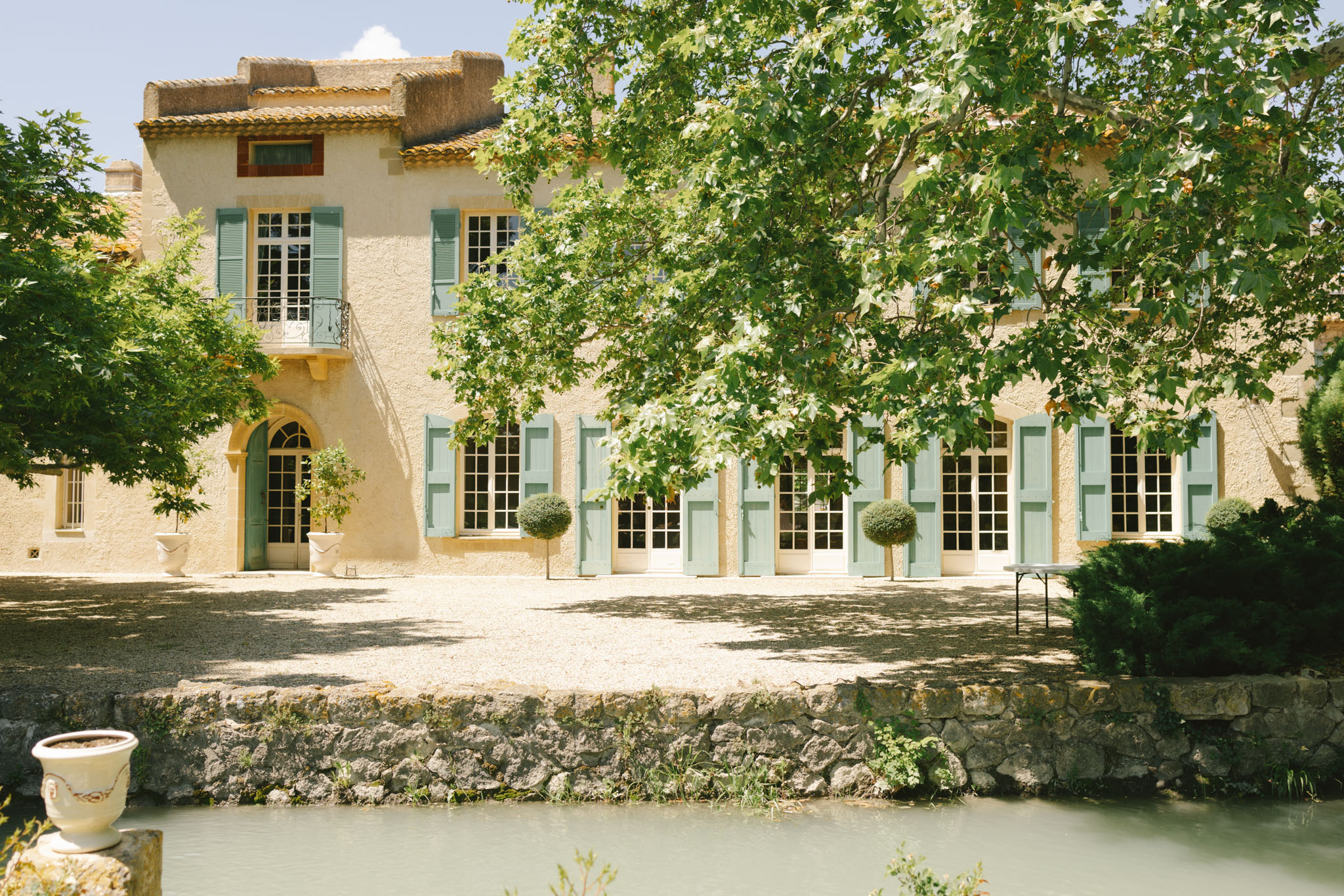 Cream stone French country house with pale blue-green shutters, ivy-covered walls, and canal in foreground