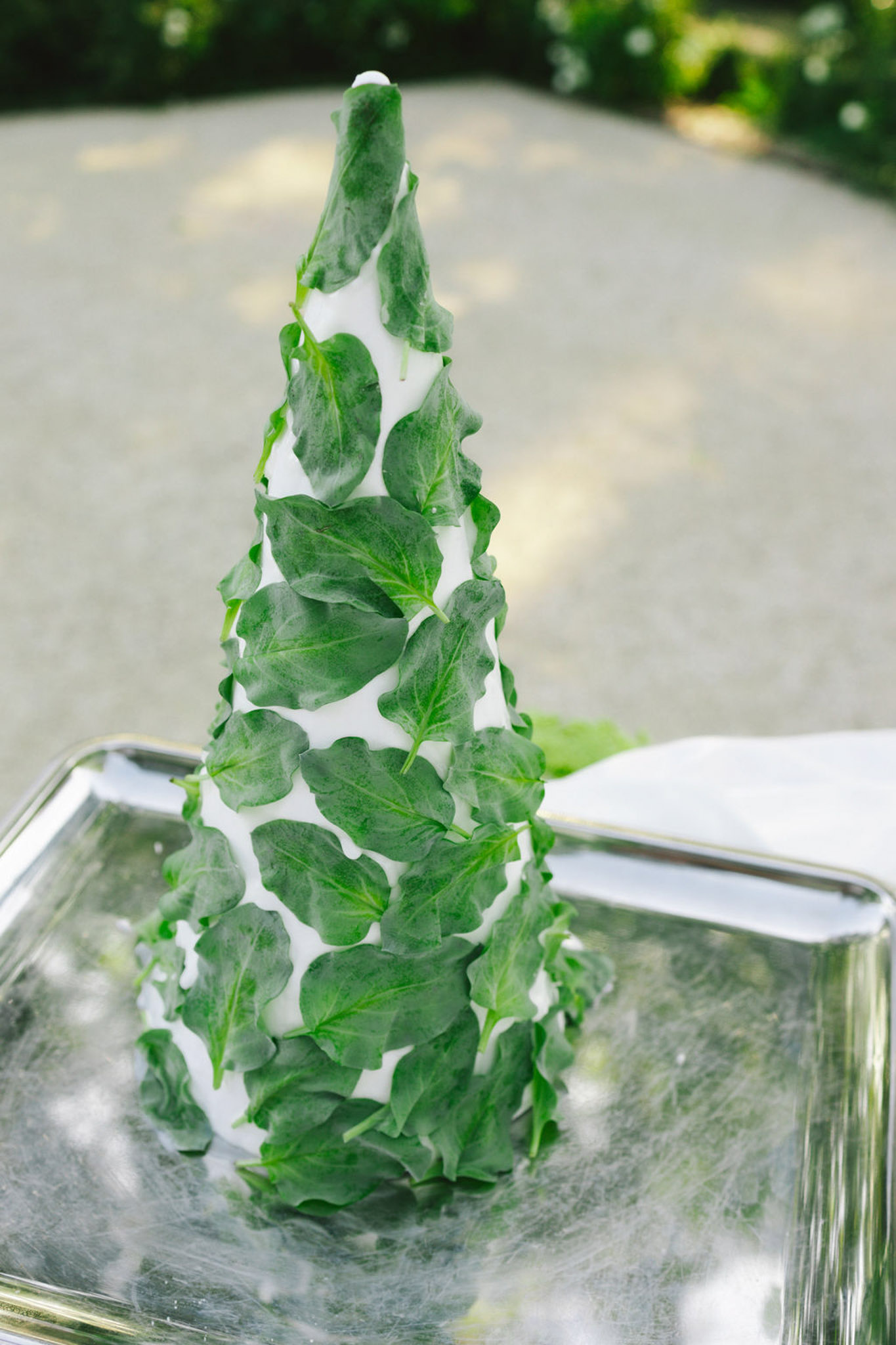 Close-up of cone-shaped green foliage centerpiece in glass vase with metallic rim, outdoor courtyard behind