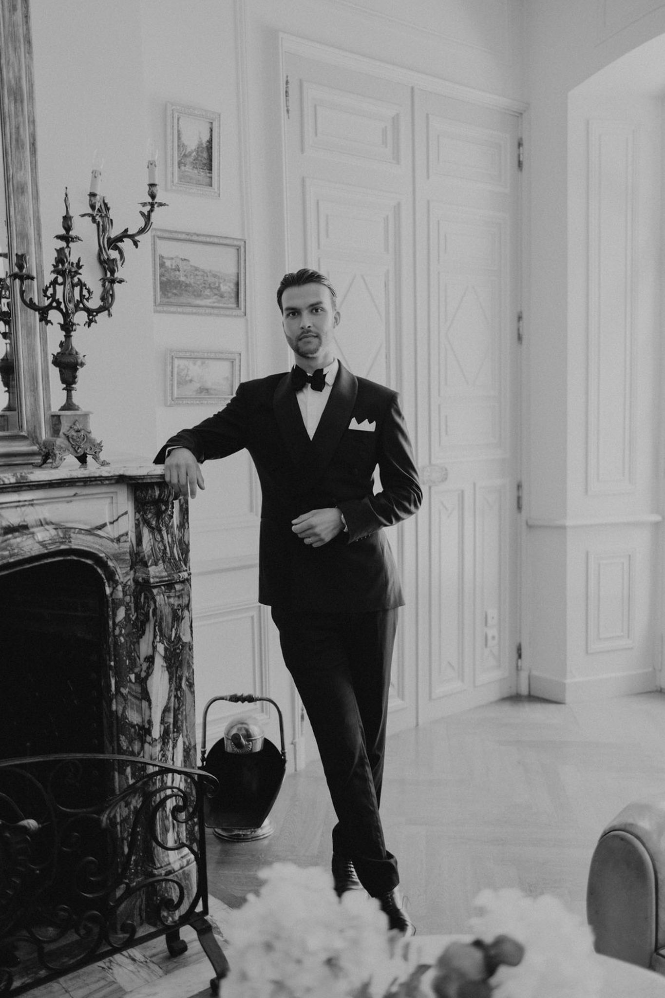 Black-and-white portrait of groom in tuxedo leaning against an ornate fireplace mantel in a classical interior