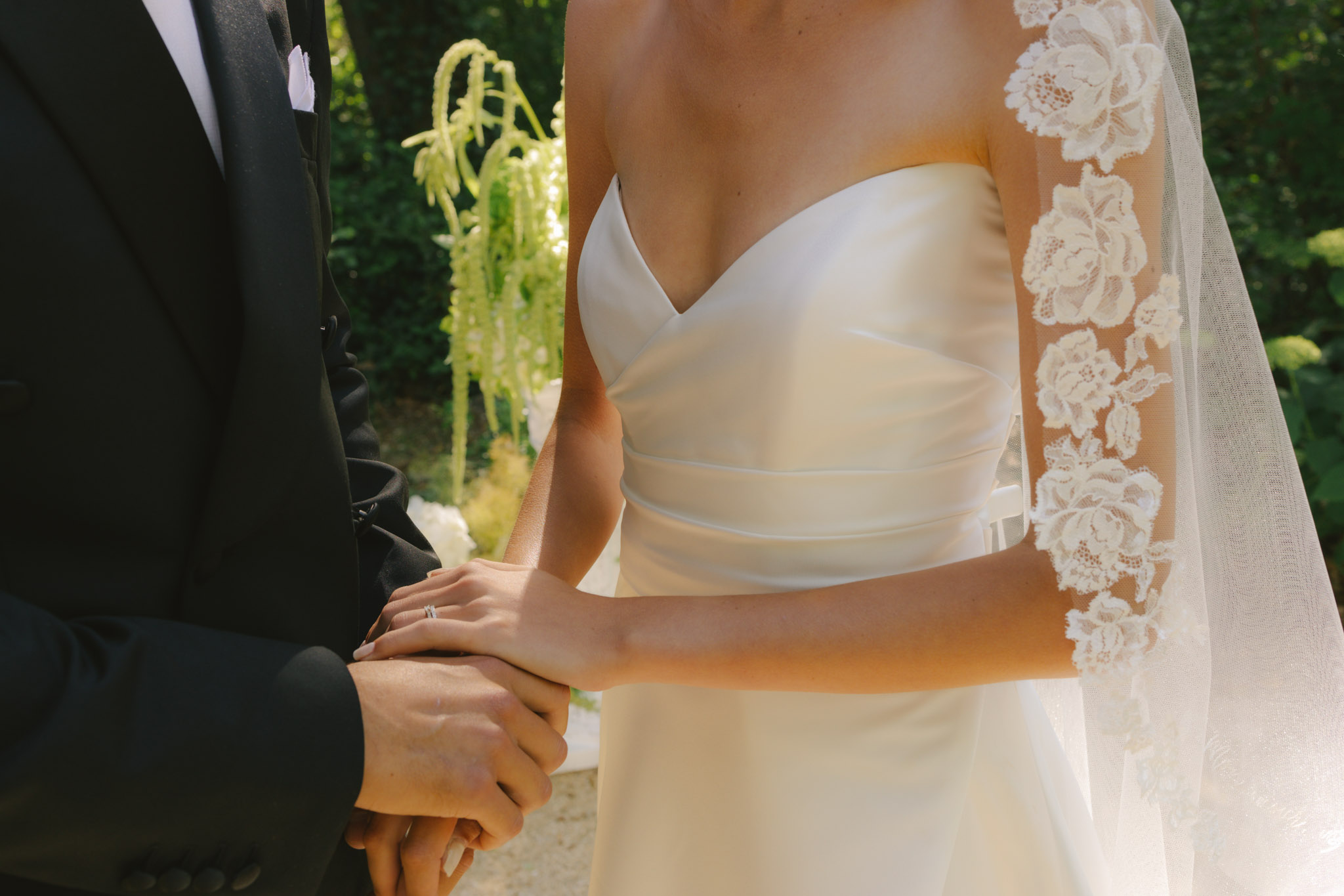 Close-up of bride and groom holding hands during ceremony, bride in lace appliqué veil and ruched ivory gown