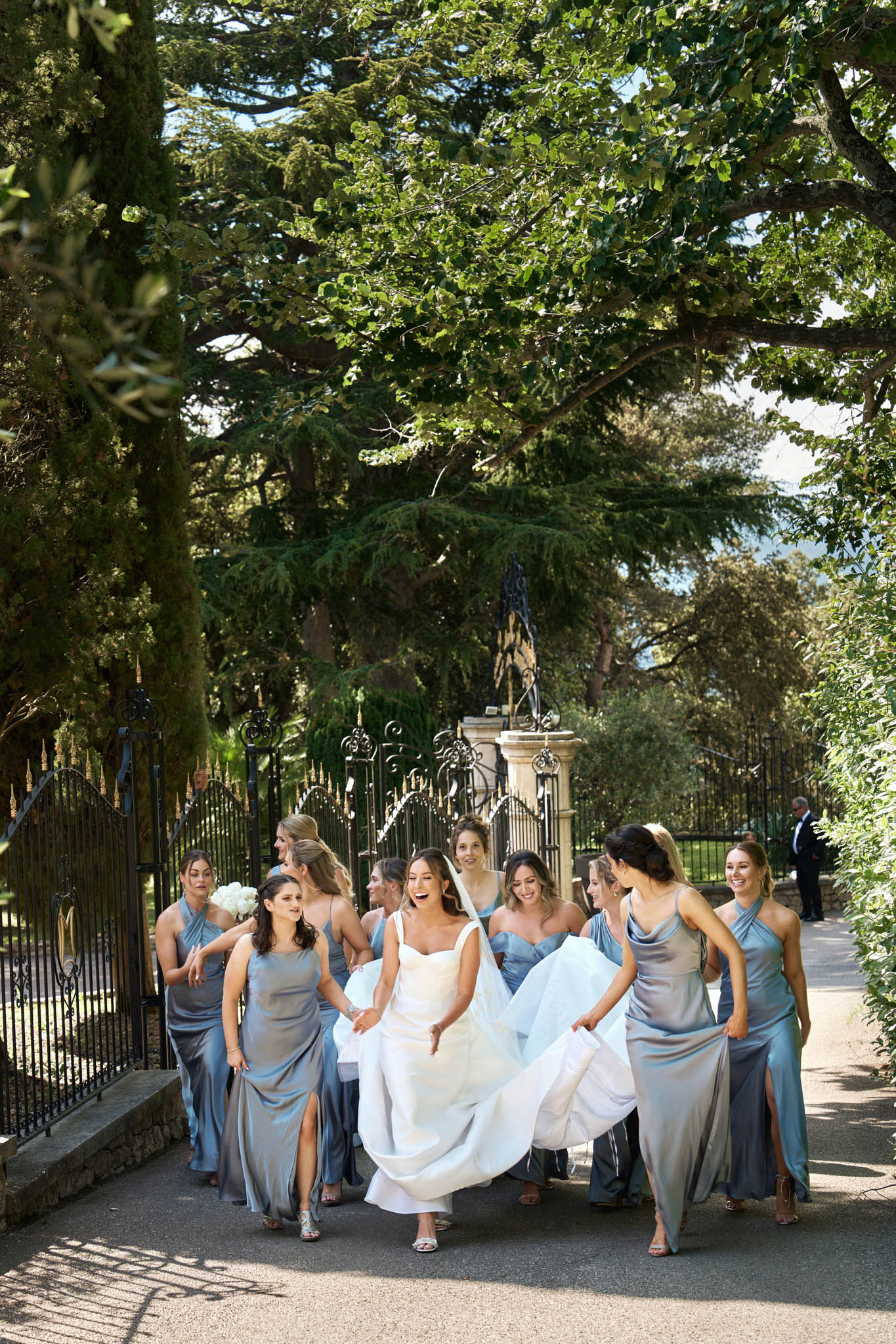 Bride and nine bridesmaids in dusty blue satin dresses walking through wrought iron gates laughing