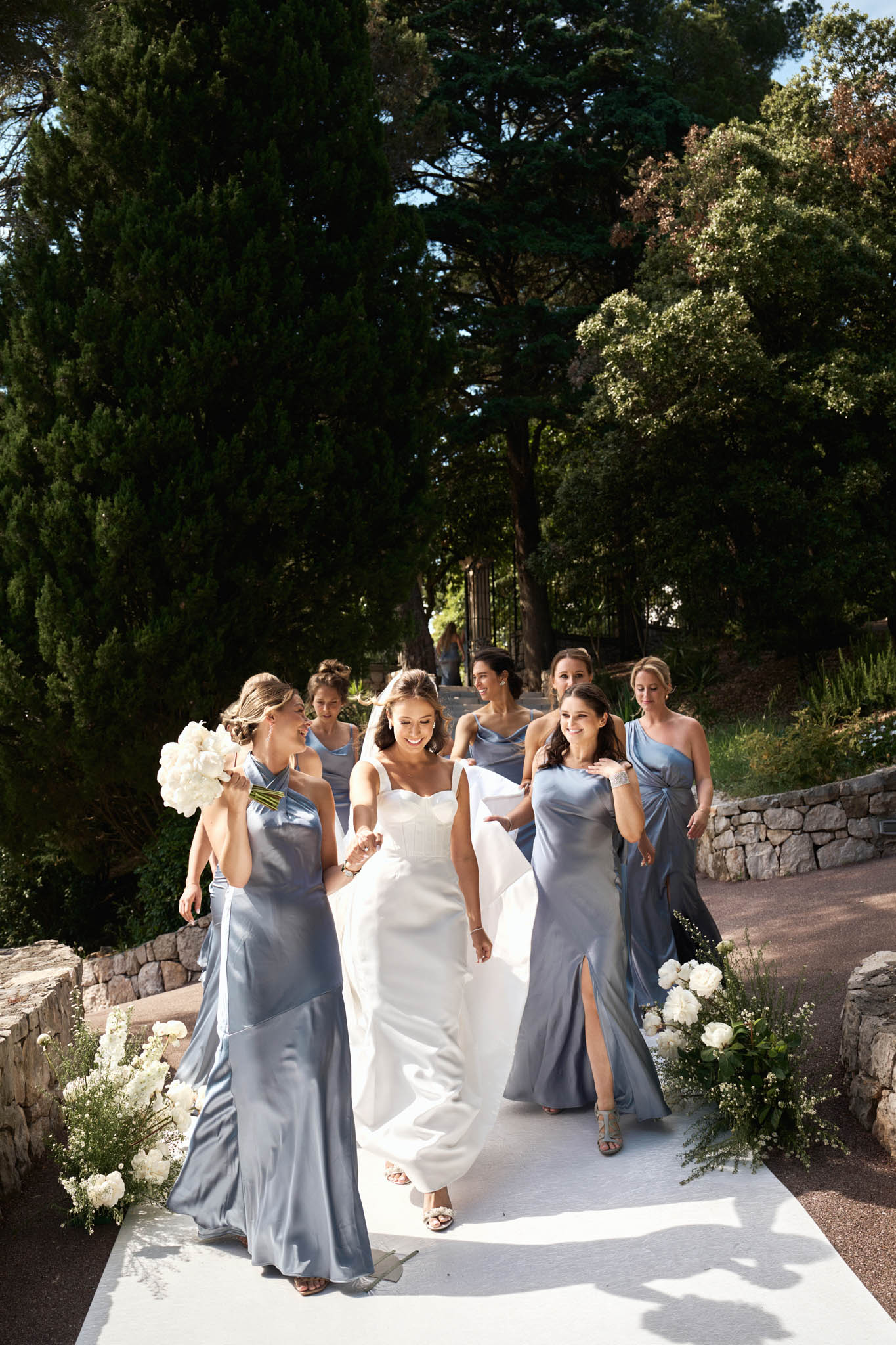 Bride in ivory bustier gown with six bridesmaids in steel-blue satin walking on white aisle with peony arrangements
