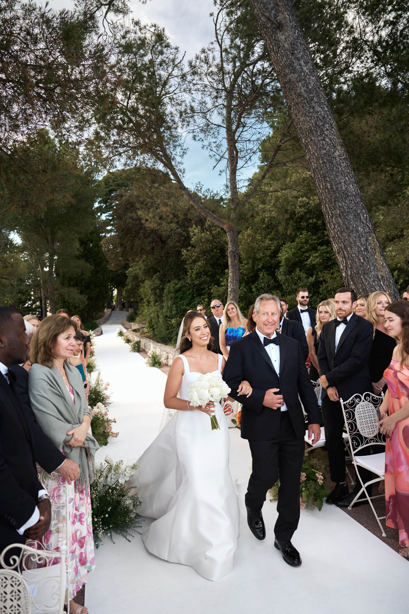 The bride is being walked down the aisle by an older man in a black tuxedo, likely her father, during an outdoor ceremony. The setting is a terrace or garden path lined with white floral arrangements featuring white roses, peonies, and greenery, with white ornate chairs seating approximately 30–40 guests on either side. The bride wears a fitted ivory sleeveless gown with a sweetheart neckline and a dramatic cathedral-length veil, and carries a round bouquet of white peonies. Guests are dressed in formal attire including black tie for some men and colorful cocktail dresses for women. The aisle is a wide white runner, and the overall styling is classic and formal. The shot is taken from a medium-wide angle slightly above eye level, capturing the full procession and the crowd of standing and seated guests reacting to the bride's entrance.