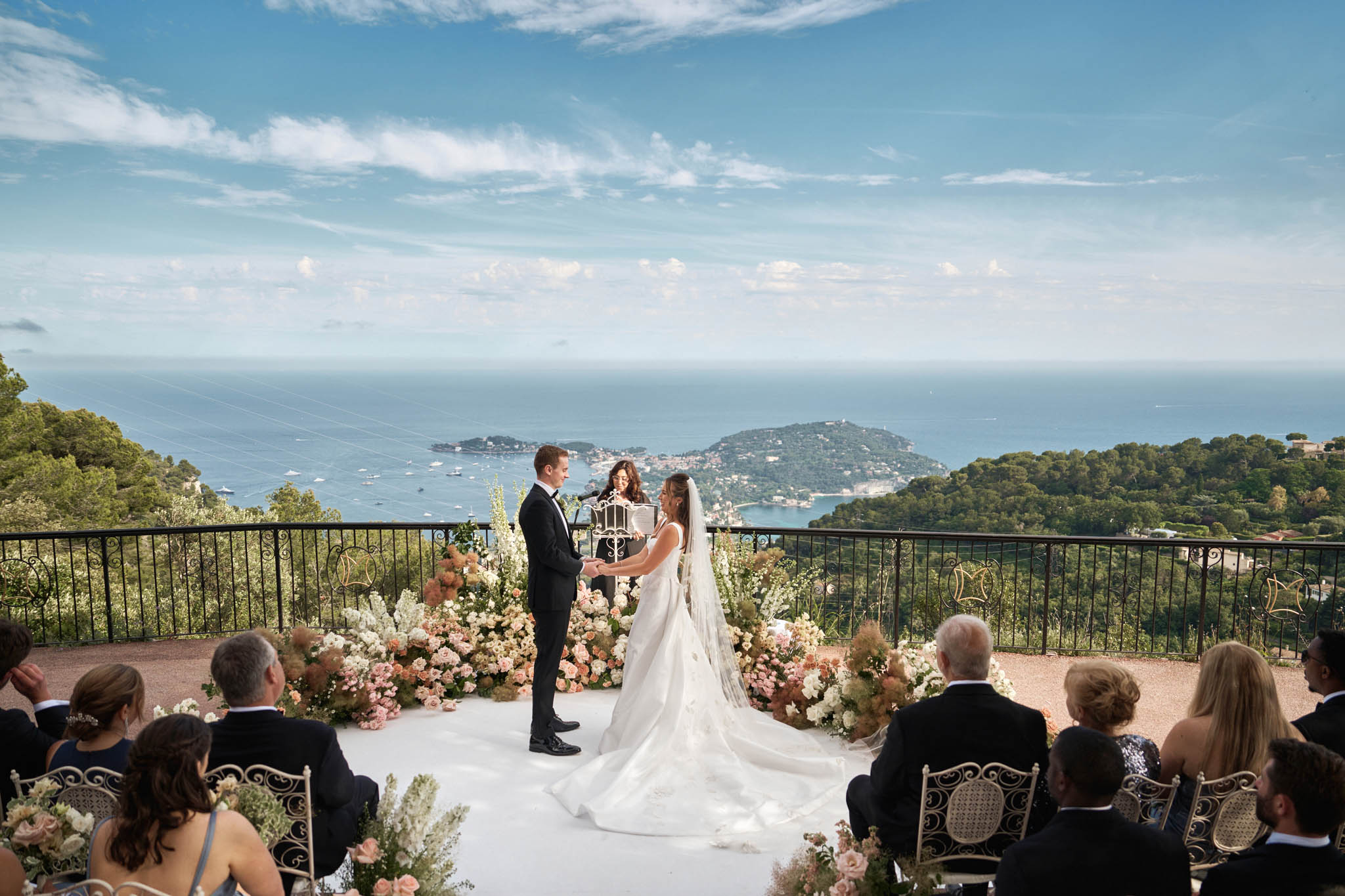 Couple on circular platform with peach and blush floral arch on terrace overlooking Mediterranean coast
