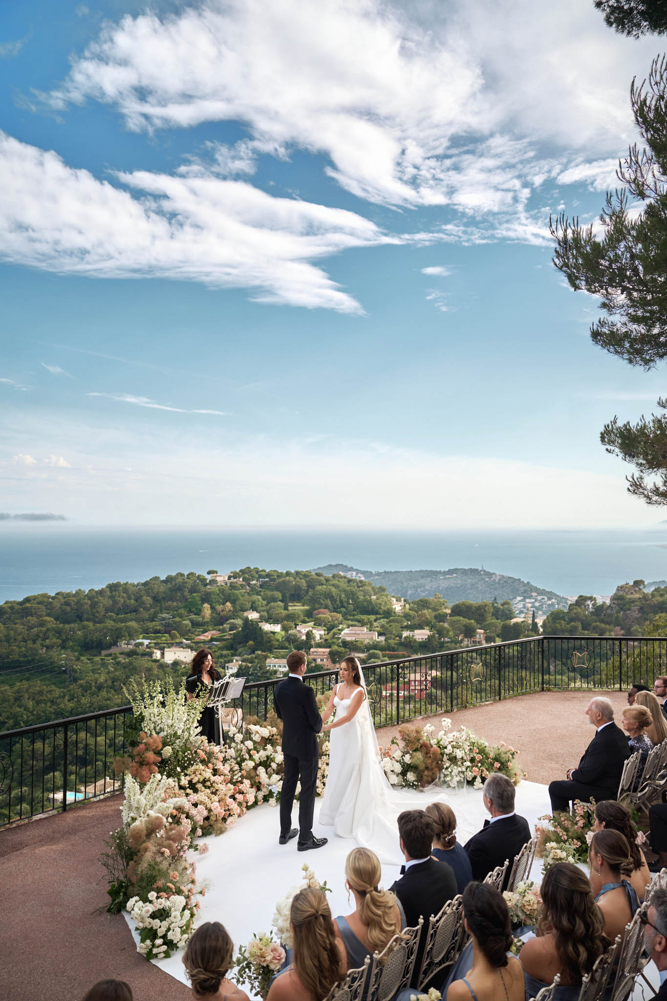An outdoor wedding ceremony taking place on an elevated terrace with a panoramic view of the Mediterranean coastline and hillside below. The bride, wearing a white sleeveless gown with a full skirt and short train, faces the groom, who is dressed in a black suit, as they exchange vows before an officiant standing at a lectern. The ceremony altar is framed by large, lush floral arrangements in blush pink, peach, ivory, and white, featuring roses, white delphinium, and pampas grass, lining a white platform stage on both sides. Approximately 30–40 seated guests, many in formal black tie attire with several women in dusty blue dresses, watch from ornate metal chairs arranged in rows on the terrace. The wide-angle shot is taken from an elevated angle behind the guests, emphasizing the dramatic Mediterranean backdrop with a black iron railing border. Potential venue feature image.
