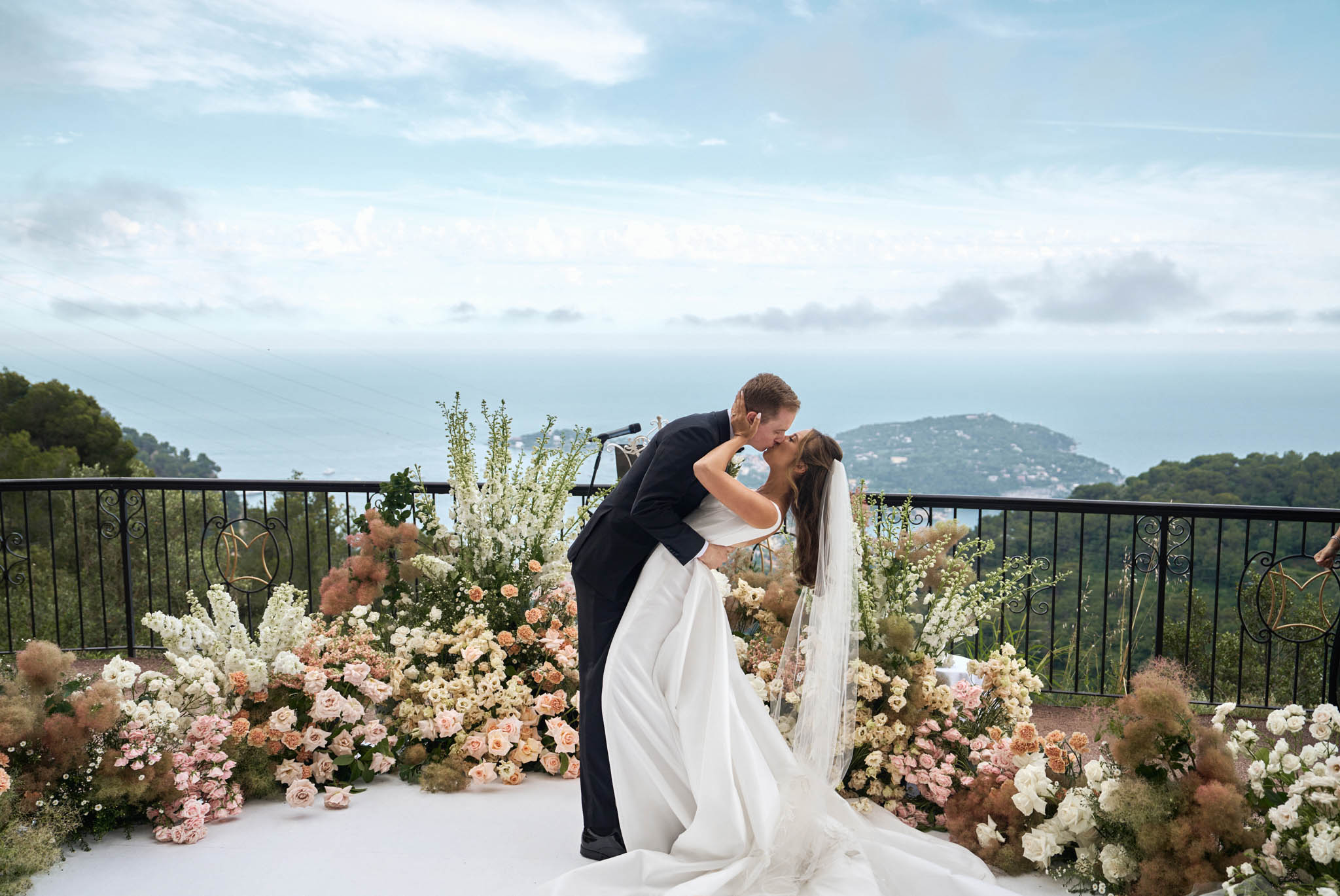 Groom dipping bride for first kiss on coastal terrace with blush rose pampas grass installation and sea panorama