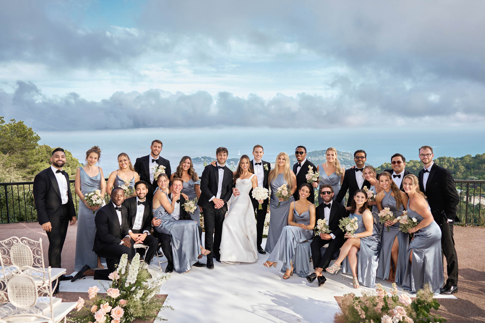 A large bridal party group portrait taken outdoors on a terrace or elevated platform with a panoramic coastal view in the background. The couple stands at the center — the bride in a fitted ivory square-neck gown holding a bouquet of white and cream blooms with greenery, and the groom in a black tuxedo with a bow tie. Approximately 20 bridal party members surround them, with bridesmaids wearing dusty blue-grey satin slip dresses in varying styles and holding bouquets of ivory and cream flowers with soft greenery, and groomsmen dressed in black tuxedos with bow ties. The foreground features blush pink and white floral arrangements at ground level, and white ornate chairs are visible to the left. The styling palette is classic and modern, combining dusty blue, ivory, and black. Wide group shot composition.