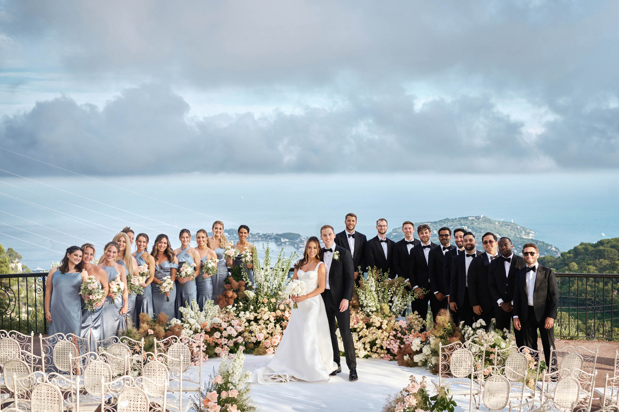 Full wedding party portrait on coastal terrace with bridesmaids in blue-grey gowns and groomsmen in black tuxedos