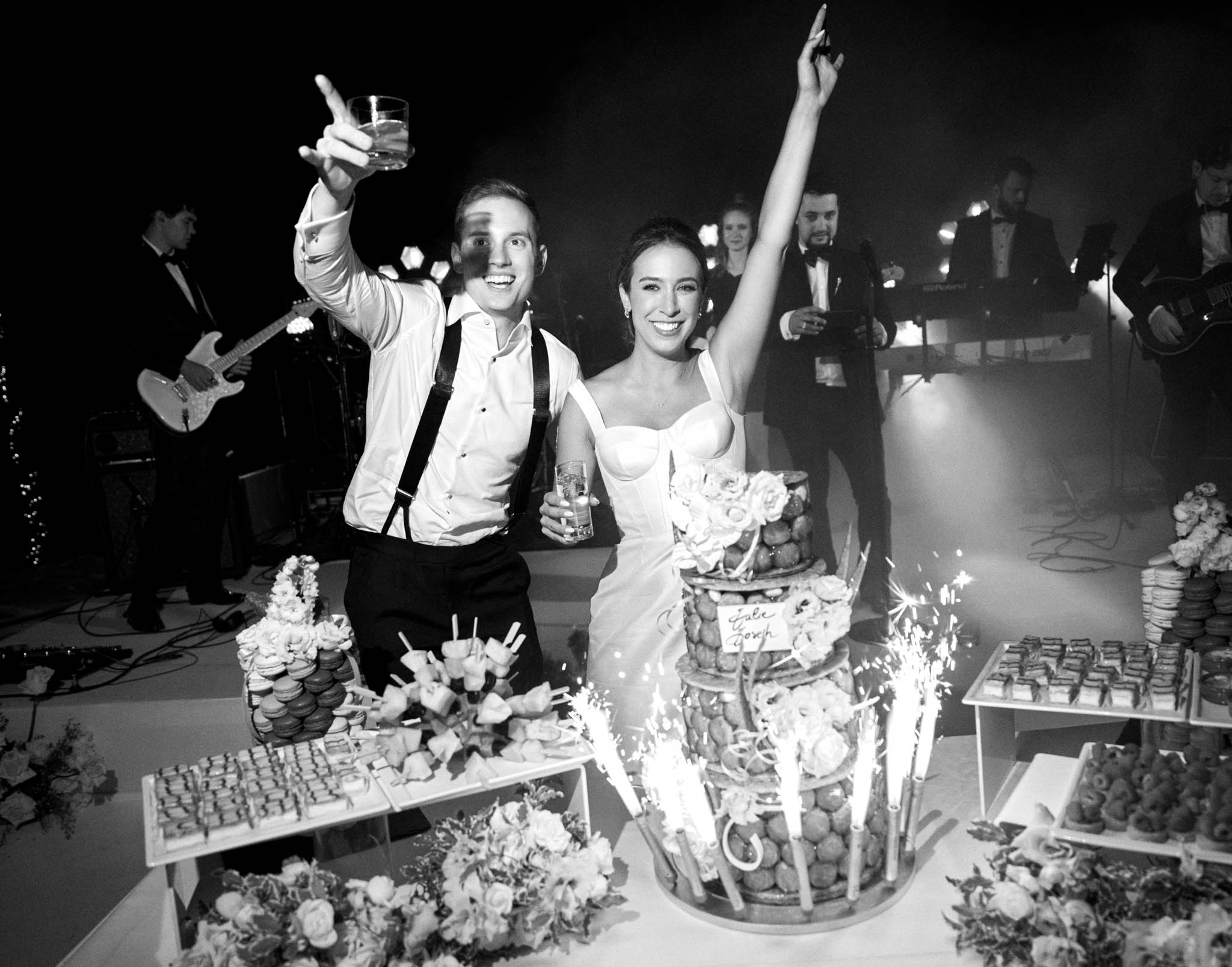 Bride and groom celebrating at croquembouche dessert table with sparklers and live band at evening reception in B&W