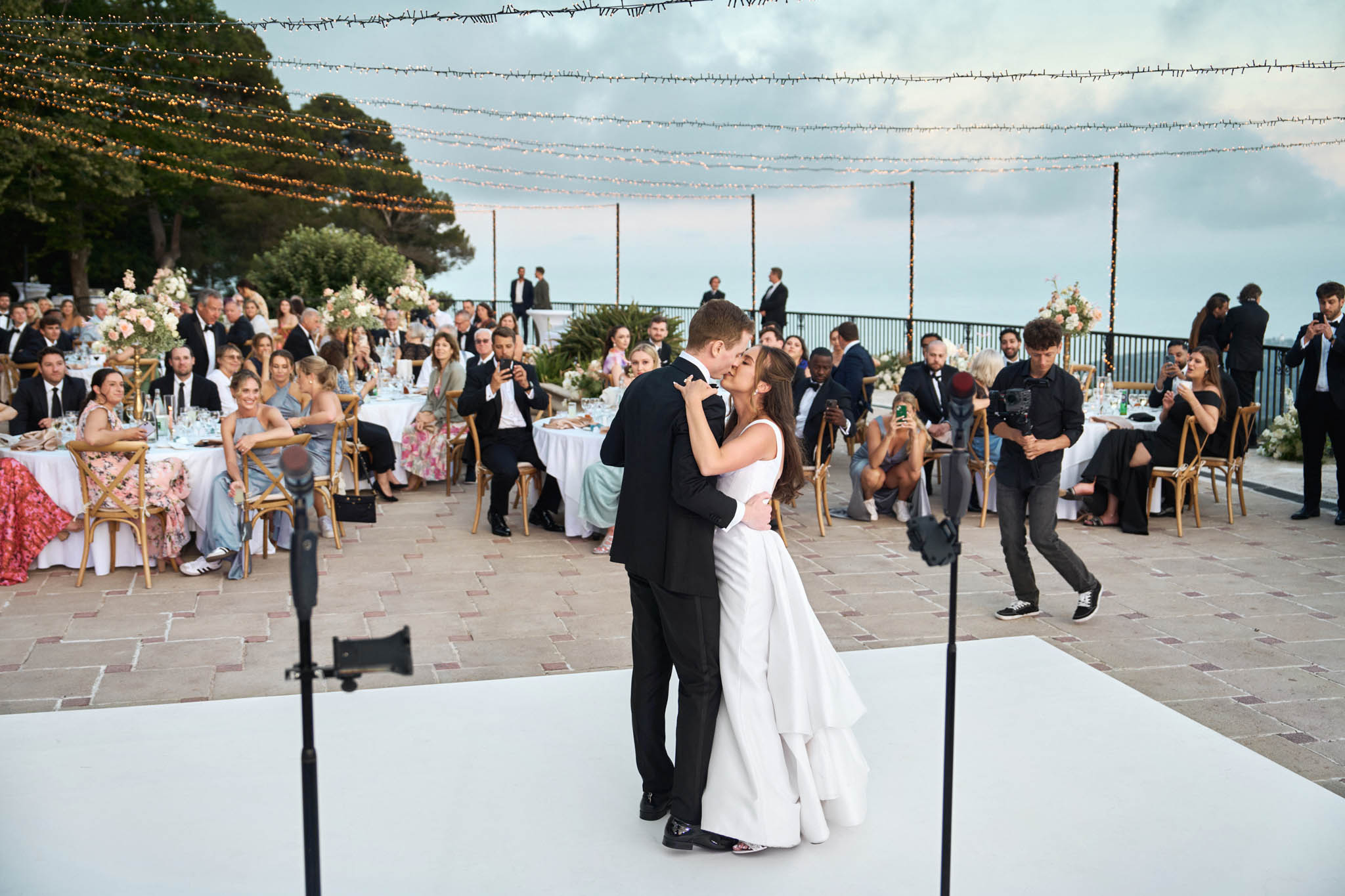 The bride and groom share a kiss during their first dance on a white rectangular dance floor at an outdoor evening reception. The bride wears a sleeveless white gown with a tiered layered skirt, and the groom is in a black tuxedo. The reception is set on a terrace with terracotta-tiled paving, surrounded by approximately 60–80 seated guests at round tables dressed in white linen, with natural wood cross-back chairs. Table centerpieces feature blush, peach, and ivory floral arrangements on tall and low arrangements. Overhead, multiple strands of warm Edison-style fairy lights and delicate floral garland lights are strung across the space on tall poles, creating a canopy effect. Several guests and a videographer are visible capturing the moment from the dance floor perimeter. The setting appears to be a clifftop or elevated coastal terrace venue, with an open-sky view and a railing along the far edge.