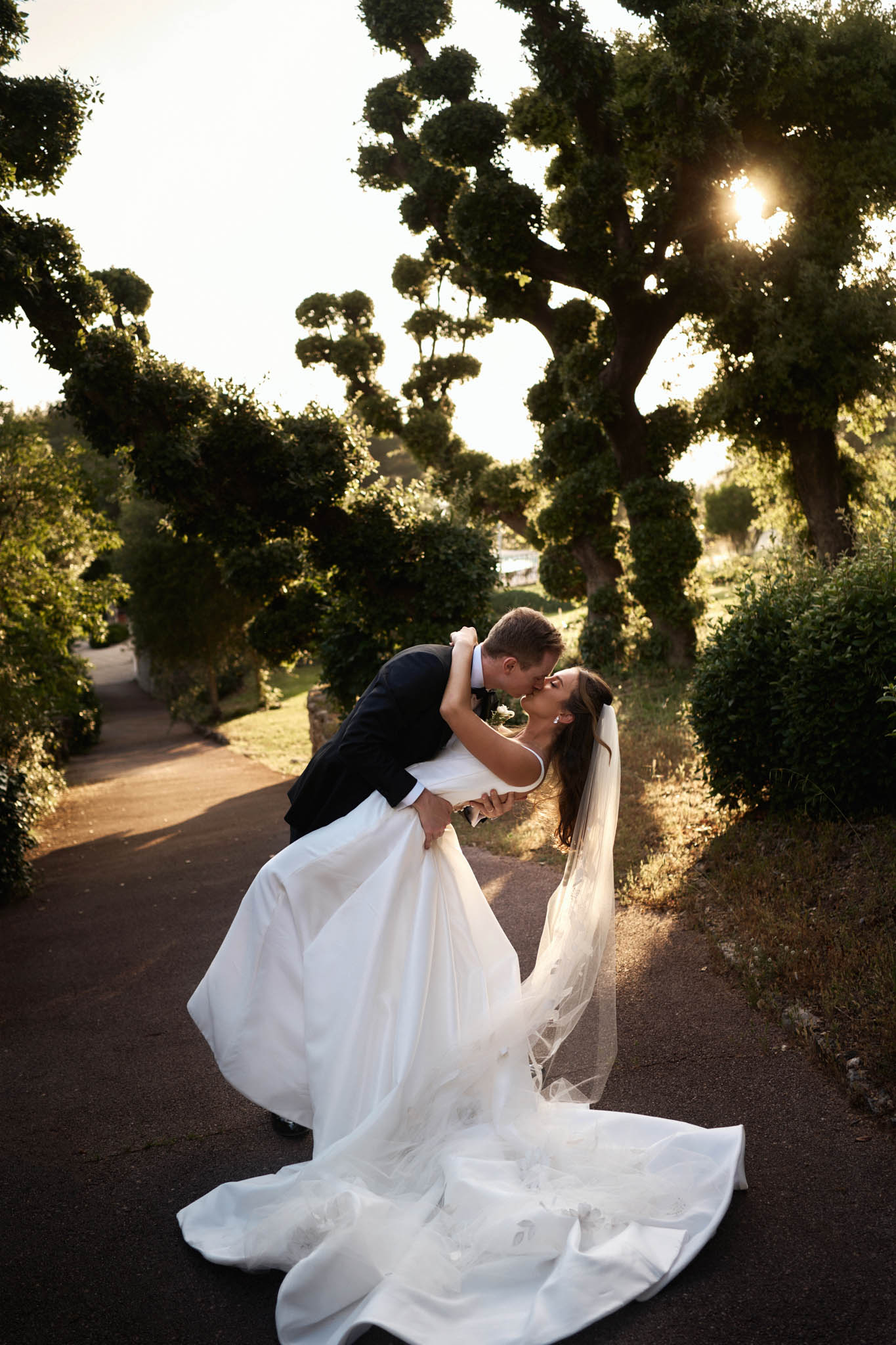 A couple portrait taken outdoors on a paved garden path lined with sculpted topiary trees. The groom, dressed in a dark navy suit, is dipping the bride toward him as they kiss. The bride wears a white off-shoulder ballgown with a long cathedral-length veil and an extended train that spreads across the path. Her dark hair is worn down. The shot is taken during golden hour, with warm backlight filtering through the tree canopy, creating a sun flare effect. The composition is a full-length portrait framed by the symmetrical tree-lined path stretching into the background.