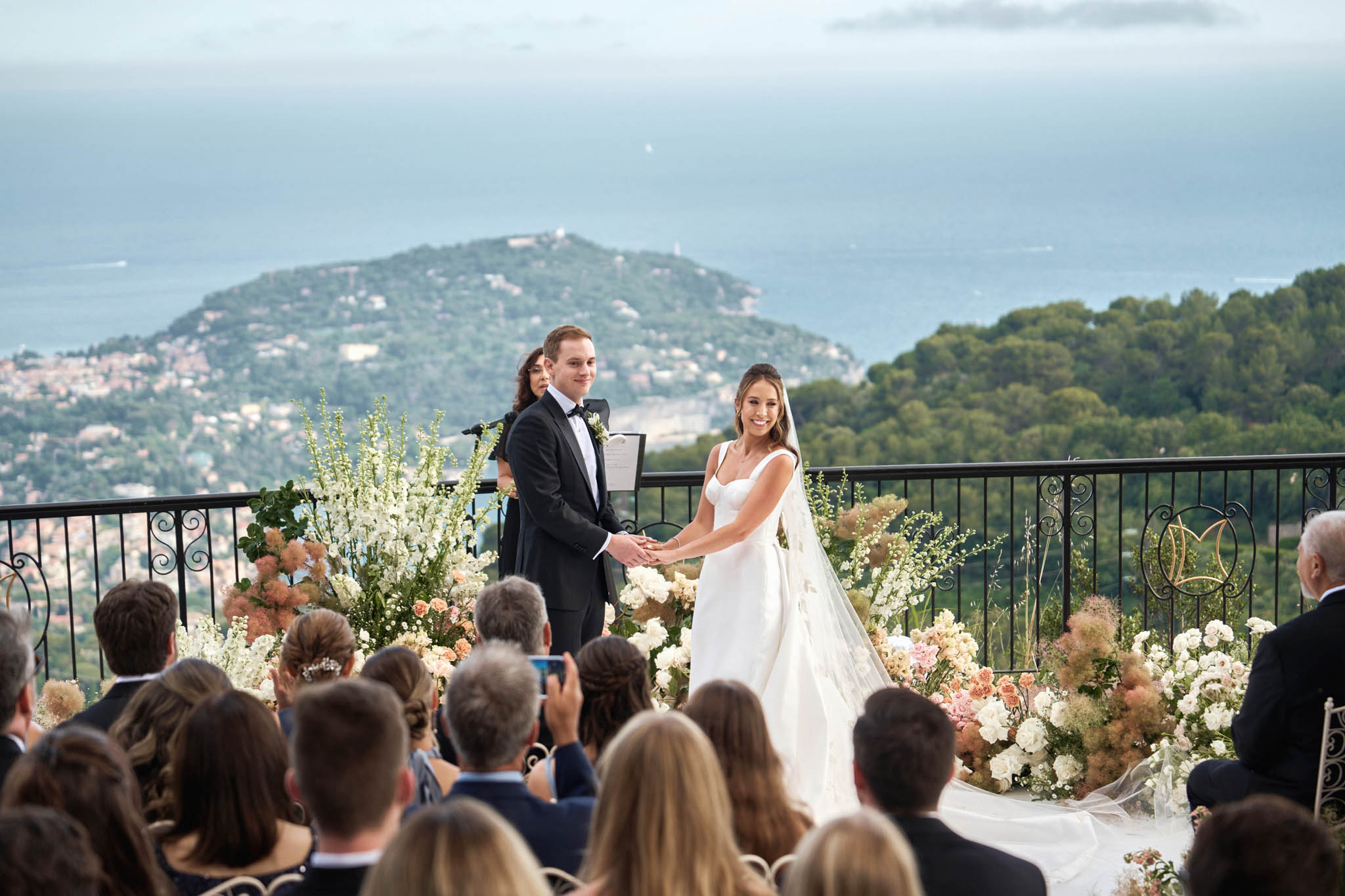 An outdoor ceremony taking place on an elevated terrace with a sweeping panoramic view of the French Riviera coastline and a hillside town in the background. The bride wears a fitted ivory gown with a structured square neckline and a long flowing veil, while the groom is dressed in a black tuxedo with a black bow tie and a white boutonniere; the couple holds hands facing their guests. A female officiant stands behind them holding a ceremony booklet, flanked by lush floral arrangements featuring white delphinium, ivory and blush roses, peach spray roses, and dried pampas-style grasses forming a ground-level floral display along the terrace railing. A gold monogram decorative element is visible on the wrought-iron railing to the right. Approximately 20–30 seated guests are visible in the foreground, and the wide-angle shot captures both the ceremony in full and the dramatic coastal backdrop. Potential venue feature image.