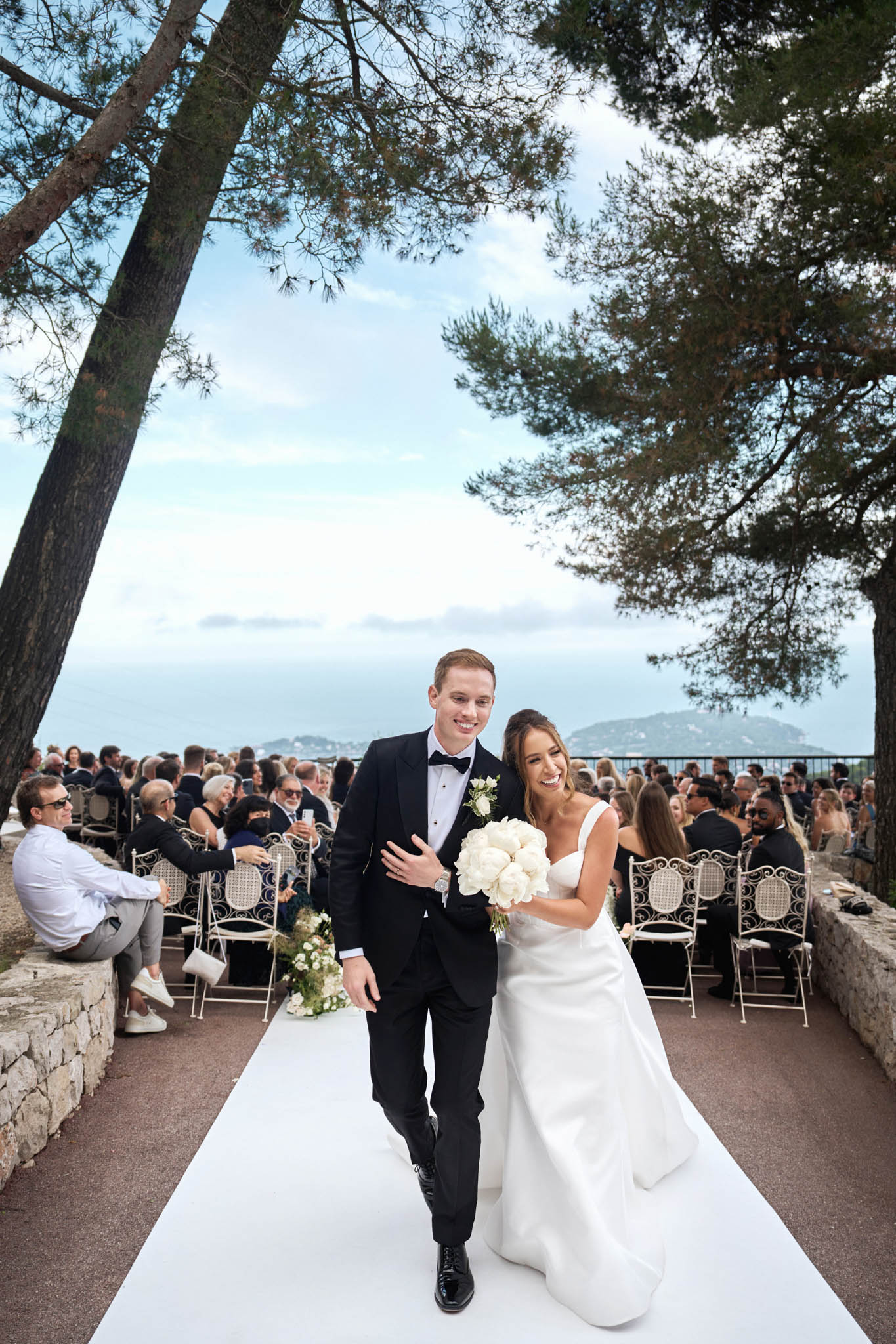 Bride and groom walk down aisle after ceremony with sea view backdrop and guests in white wrought-iron chairs