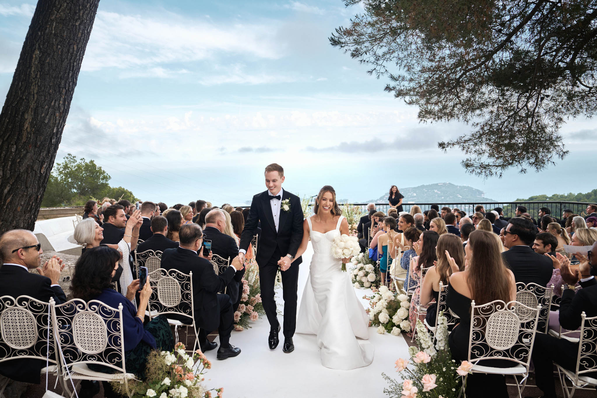The bride and groom walk back up the aisle together following the conclusion of an outdoor ceremony, both smiling and holding hands. The ceremony takes place on a terrace with a sea view and distant hills visible in the background, suggesting a Mediterranean coastal venue. The groom wears a black tuxedo with a white boutonnière, while the bride wears a fitted ivory structured gown with a sweetheart neckline and carries a full bouquet of white peonies. The aisle runner is white and lined on both sides with low floral arrangements of white peonies, blush and peach roses, and greenery; seating consists of ornate white wrought-iron chairs filled with approximately 80–100 guests who are applauding. An officiant stands at the far end of the aisle near a railing overlooking the sea. The overall decor palette is white, blush, and peach with a classic, refined outdoor ceremony style. Wide shot taken from a low angle looking up the aisle toward the couple.
