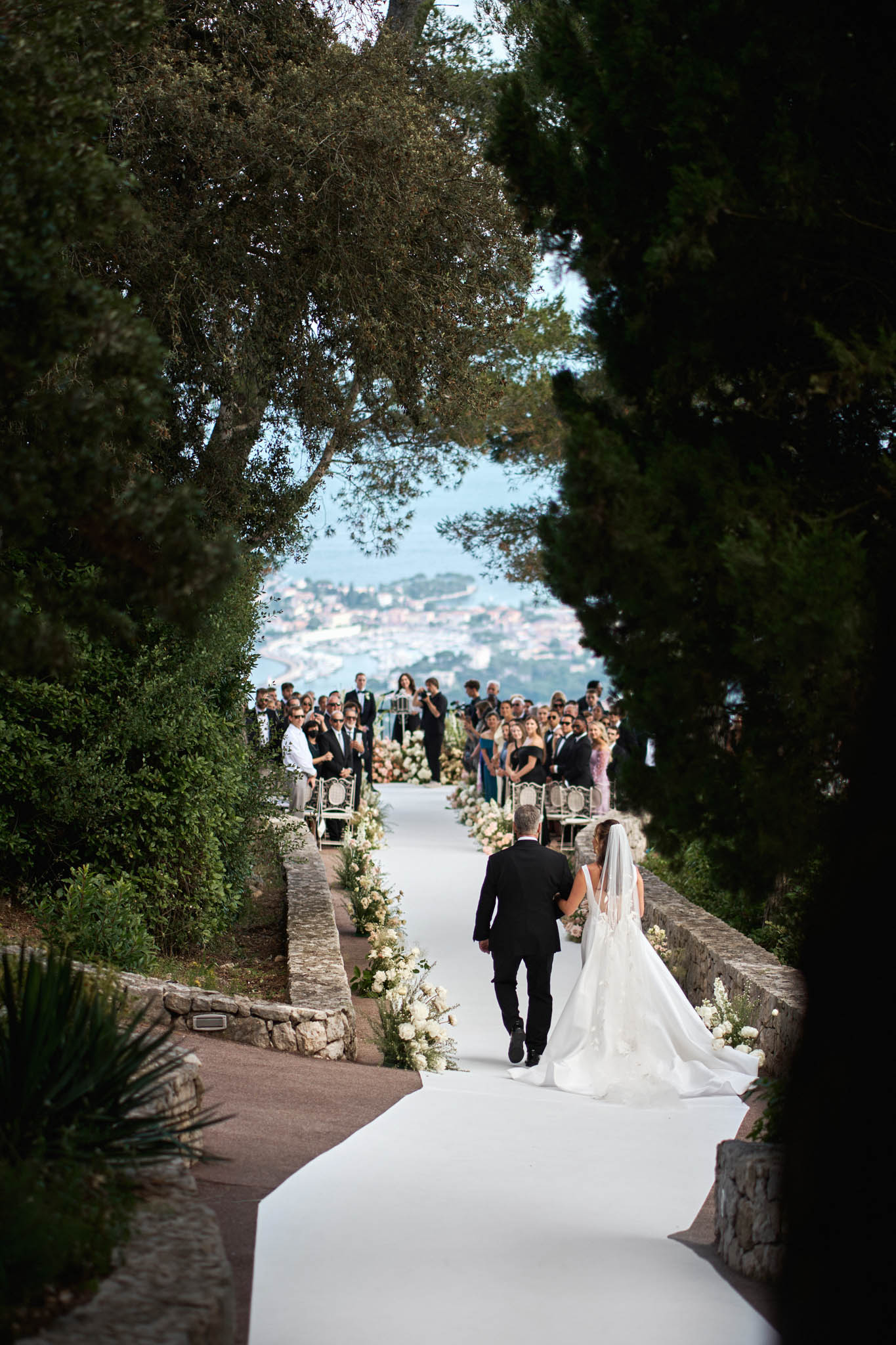 Bride walking down flower-lined aisle between cypress trees with coastal panorama at hilltop Riviera venue