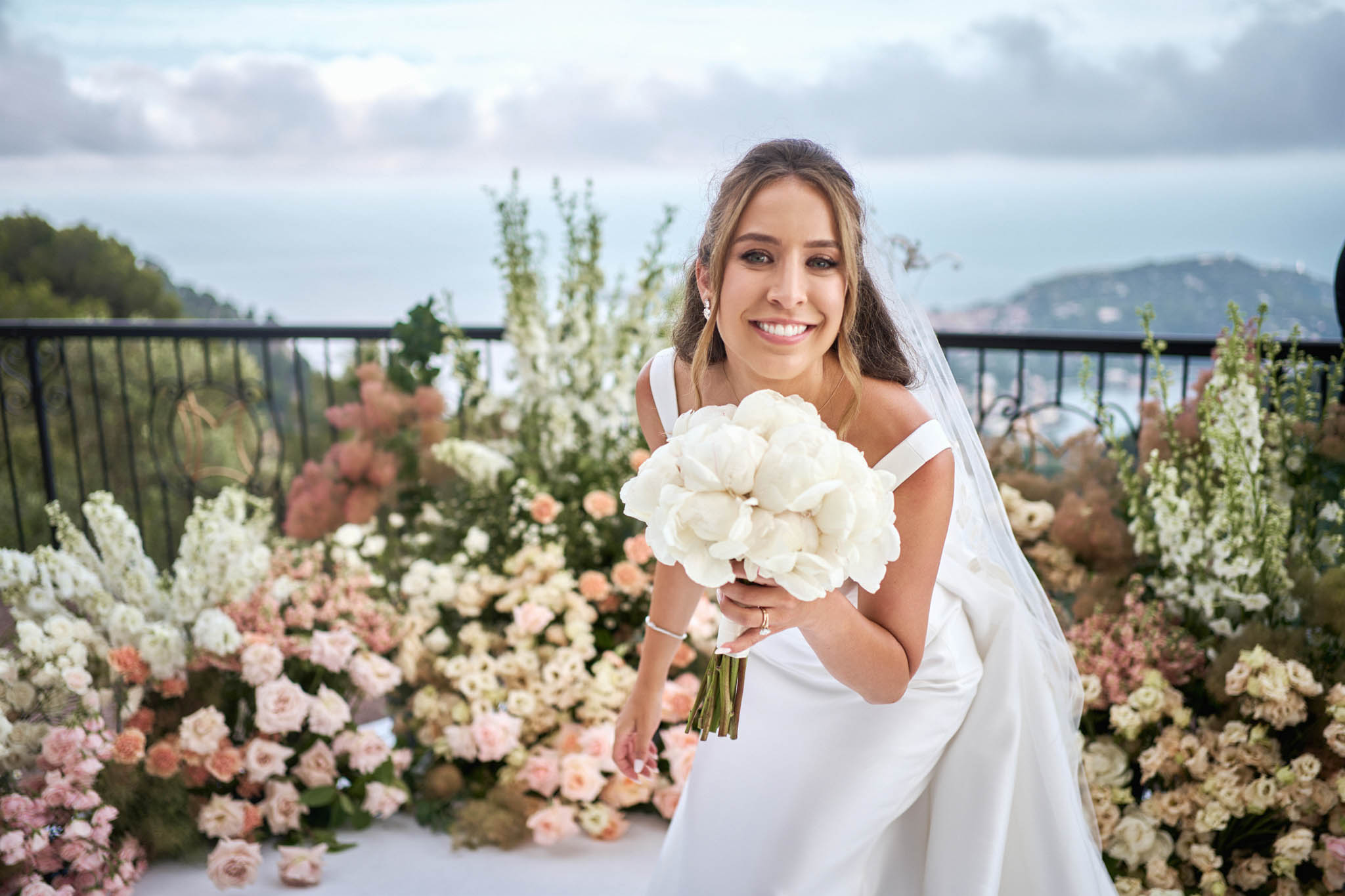 A bridal portrait taken outdoors on a terrace or balcony with a wrought-iron railing and a hillside and water view in the background. The bride wears an off-the-shoulder white satin gown with a cathedral veil and holds a tightly arranged bouquet of white peonies with green stems. She is leaning slightly forward toward the camera and smiling directly at the lens. Behind her is a large ground-level floral installation featuring blush pink roses, peach spray roses, white delphiniums, white snapdragons, and dusty pink blooms arranged in a lush, garden-style display. The overall decor palette is white, blush, and peach with greenery accents, styled in a romantic, classic aesthetic. Medium close-up portrait composition.