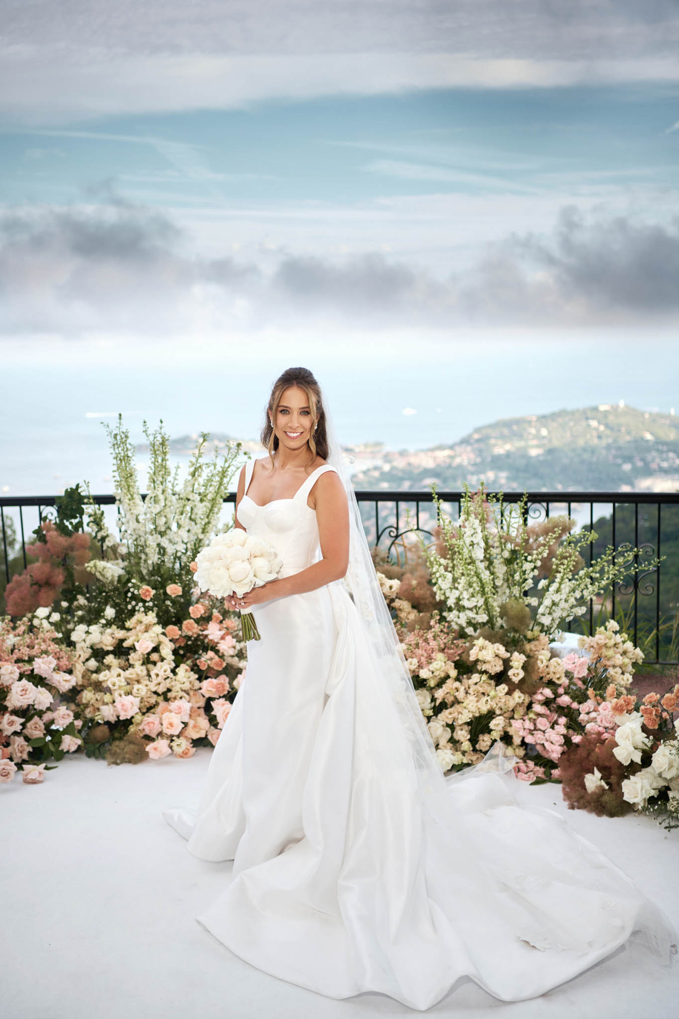A bridal portrait taken outdoors on a terrace or ceremony platform with a panoramic coastal view visible behind a black iron railing. The bride stands alone in a white ball gown with a structured sweetheart bodice, wide straps, and a long cathedral train, paired with a cathedral-length veil. She holds a rounded bouquet of white peonies and carries a warm, confident expression. Behind her is a large ground-level floral arrangement featuring blush and peach roses, white delphiniums, dusty mauve smoke bush, ivory garden roses, and soft cream blooms spread across the full width of the frame in a loose, organic style. The overall floral palette is blush, peach, ivory, and mauve against a white ceremony floor. The shot is a full-length portrait framing the bride centrally against the floral backdrop and coastal scenery.