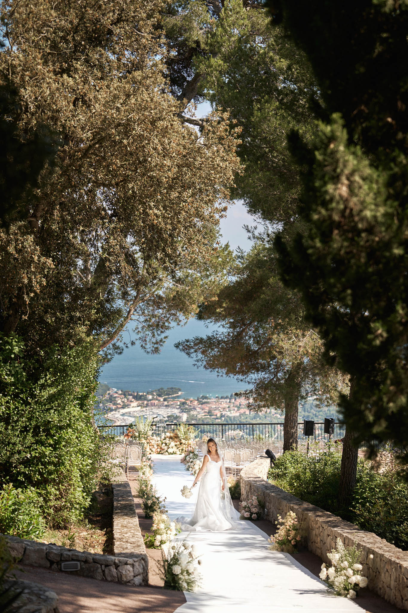 A bride in a white A-line gown with a flowing veil walks along a white aisle runner toward an outdoor ceremony space set on a terraced hillside venue overlooking a coastal town and the Mediterranean Sea. The ceremony setup includes rows of clear ghost chairs arranged on either side of the aisle, with lush floral arrangements in white, ivory, and soft blush tones lining the path and framing the altar area. The wide shot is framed by mature pine and cypress trees that create a natural archway, with the coastline and blue water visible in the background beyond a black iron railing. The overall styling is classic and garden-inspired, with an organic, softly romantic floral palette. Potential venue feature image.