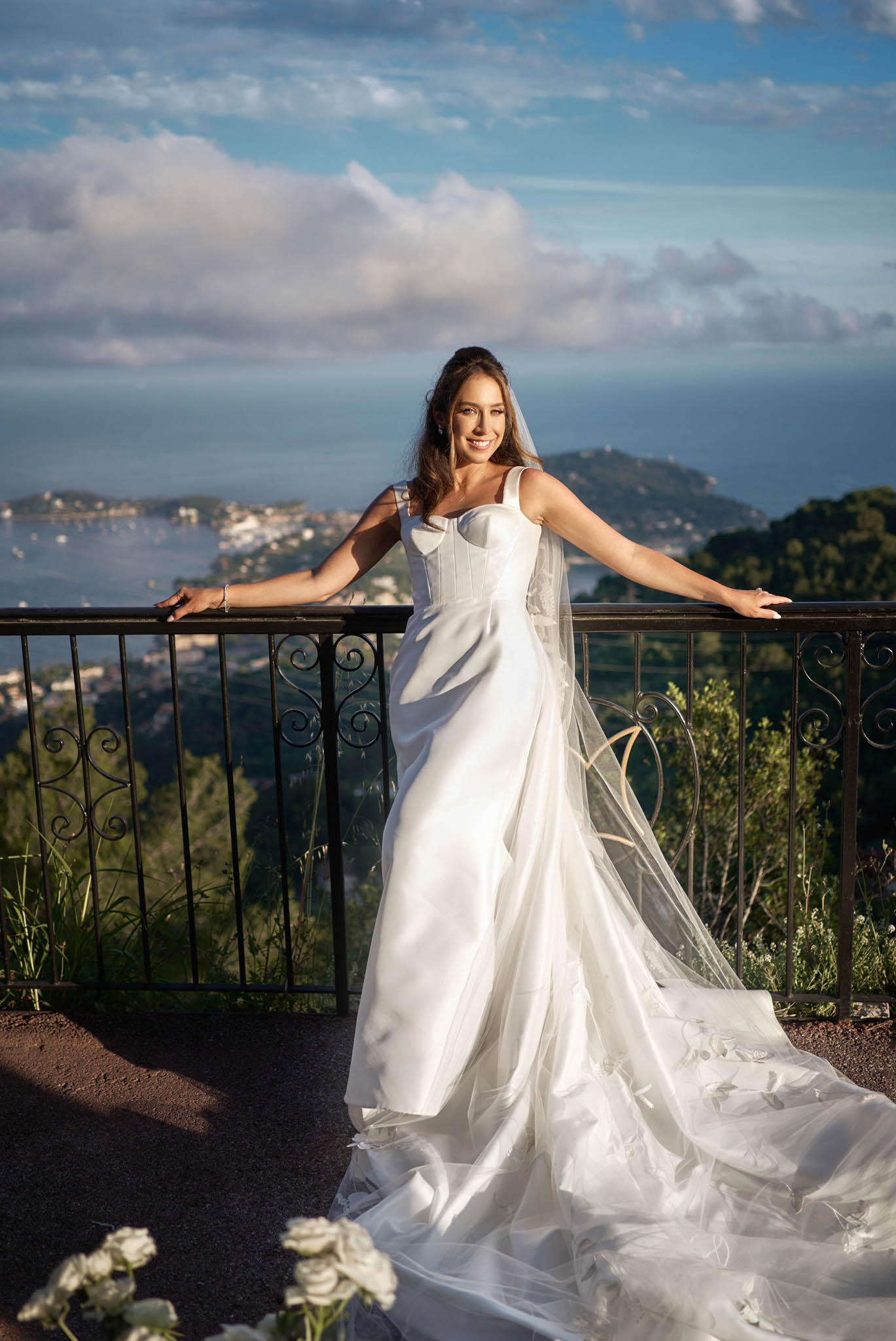 Bride in white satin gown with cathedral veil posing on wrought-iron terrace overlooking coastal bay