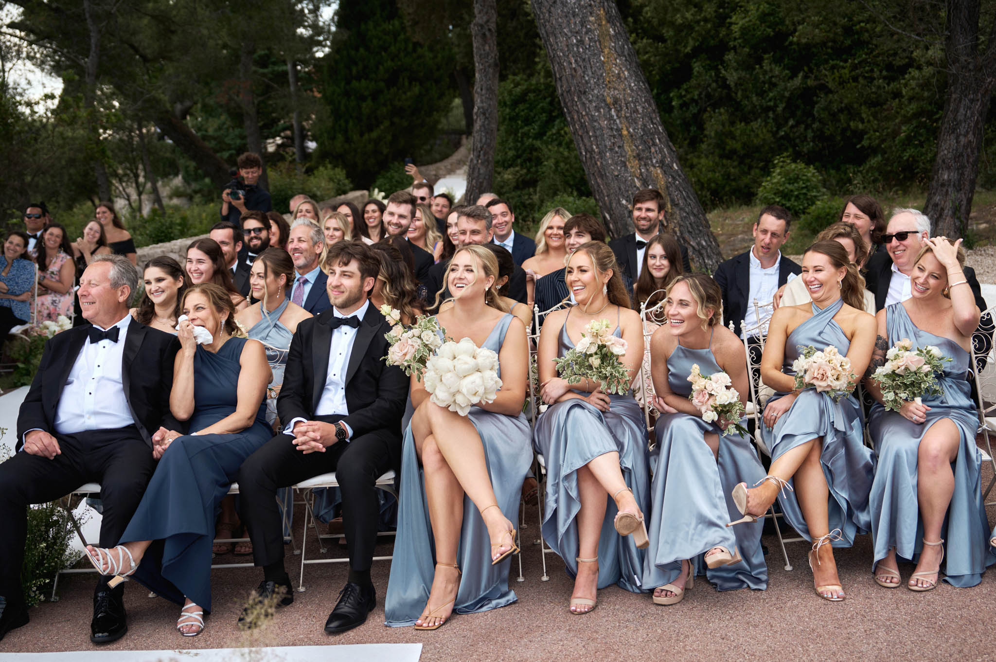 Outdoor ceremony front rows with bridesmaids in dusty blue satin dresses holding ivory and blush bouquets