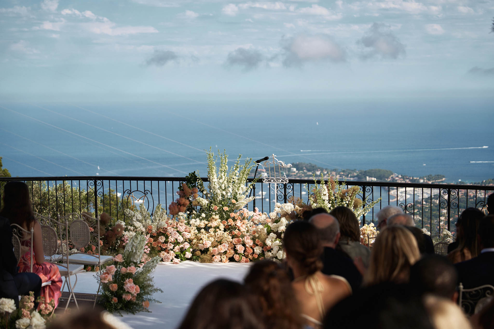 An outdoor wedding ceremony taking place on an elevated terrace with a panoramic view of the Mediterranean coastline and a coastal town visible far below. Guests are seated in rows facing a ceremony altar area framed by an abundant ground-level floral installation featuring blush pink, peach, coral, and ivory roses, white delphiniums, and soft caramel-toned dried or pampas-style blooms. Two ornate cream-colored rattan-style chairs are positioned at the altar, and a microphone stand is visible at the center. The terrace is bordered by a black wrought-iron railing with scrollwork detailing. The wide-shot composition is taken from behind the seated guests, placing the floral display and sea view as the focal point. The overall decor palette is soft and romantic, combining blush, peach, and ivory florals in a lush, garden-style arrangement.