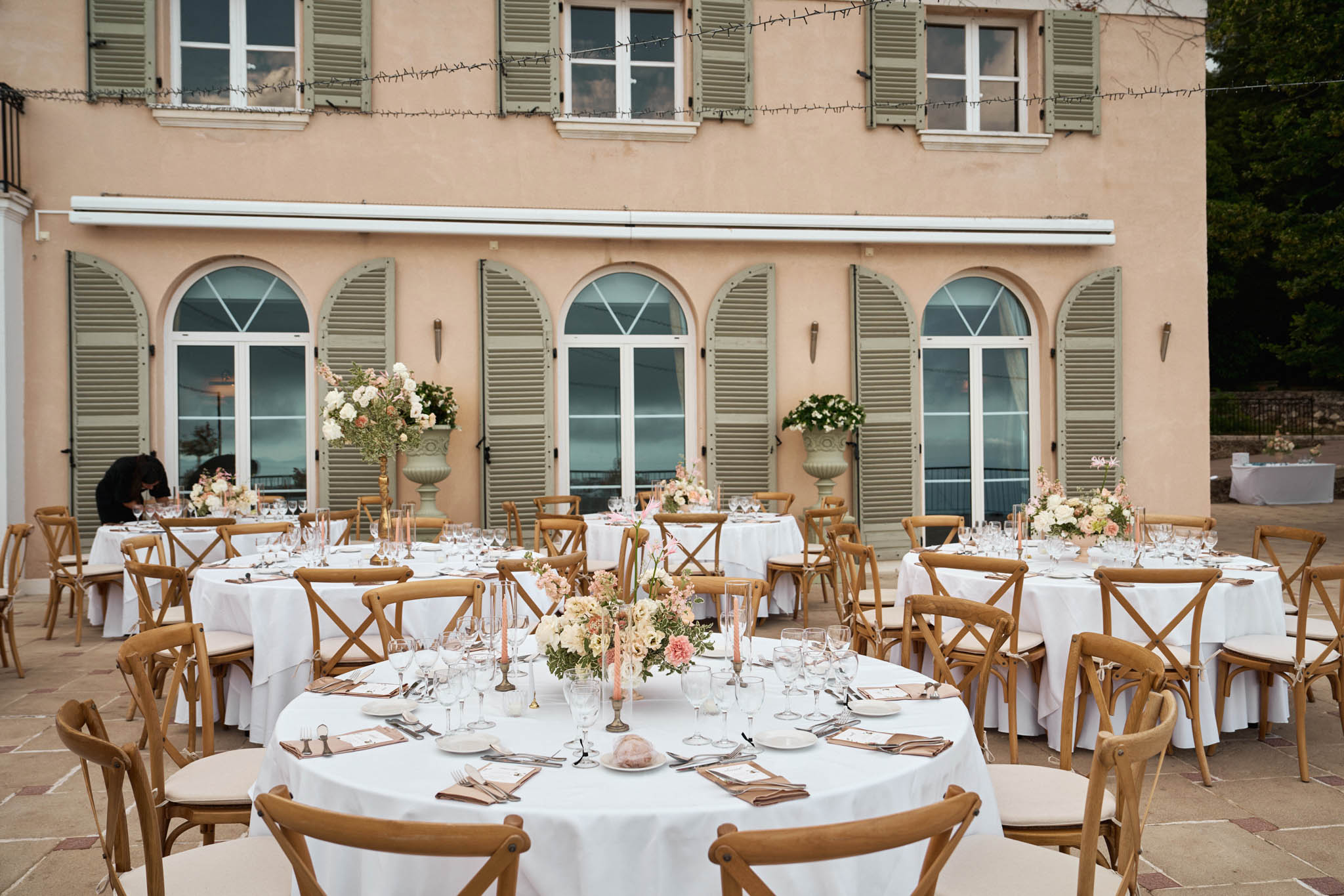 Outdoor reception tables with blush and coral centerpieces on stone terrace in front of peach French manor house