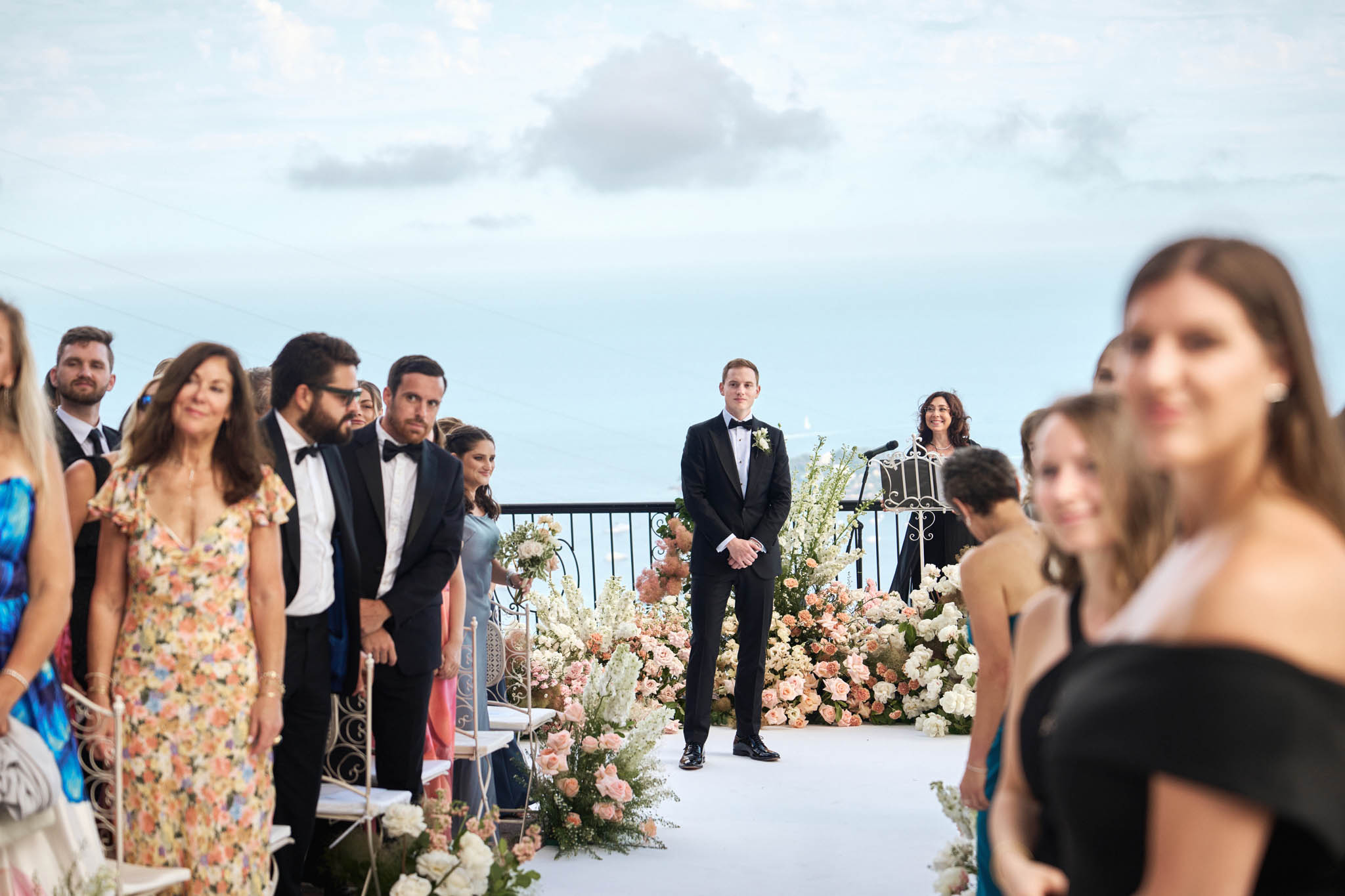 Groom waiting at coastal clifftop altar with blush and peach floral aisle arrangements guests standing on both sides