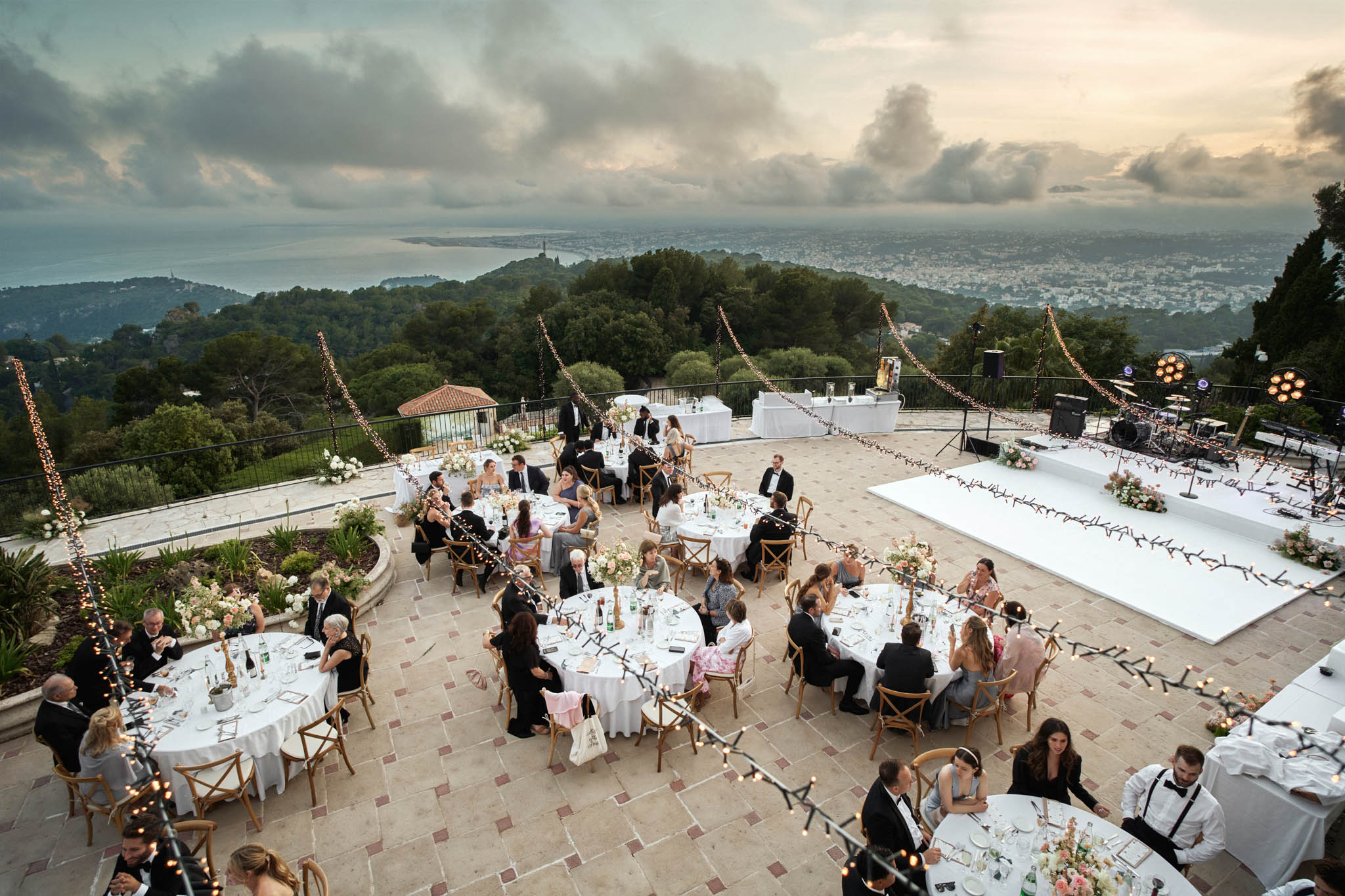 Aerial view of terrace reception with 70 guests, dance floor, and coastal city views at dusk