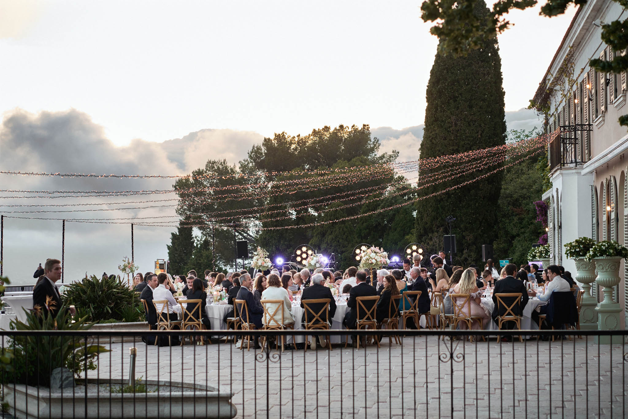 Round tables with blush centerpieces under fairy-light canopy and band stage at villa terrace at dusk