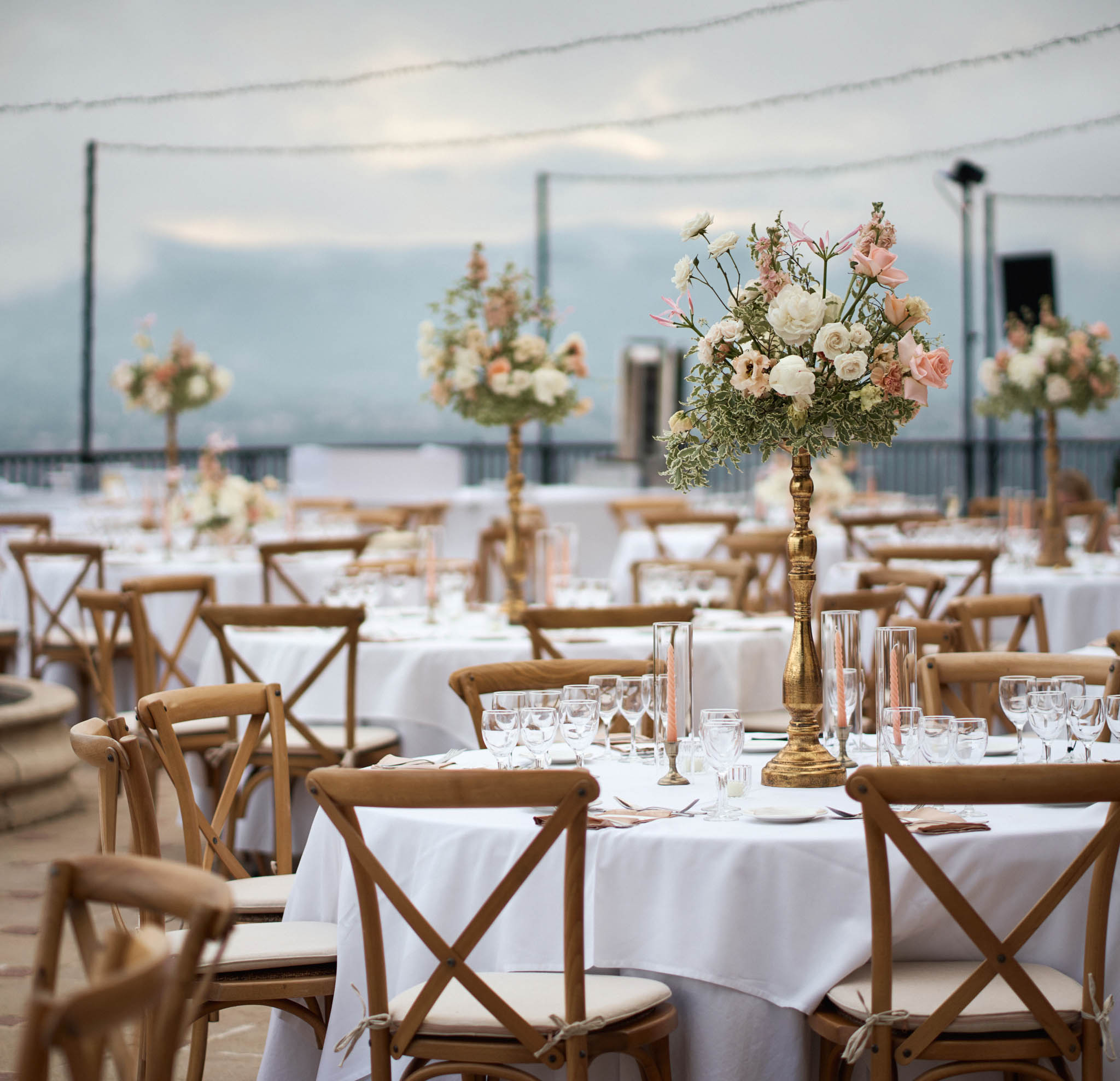 Outdoor reception with round tables, tall gold pedestal centerpieces of blush roses and ranunculus, fairy lights overhead