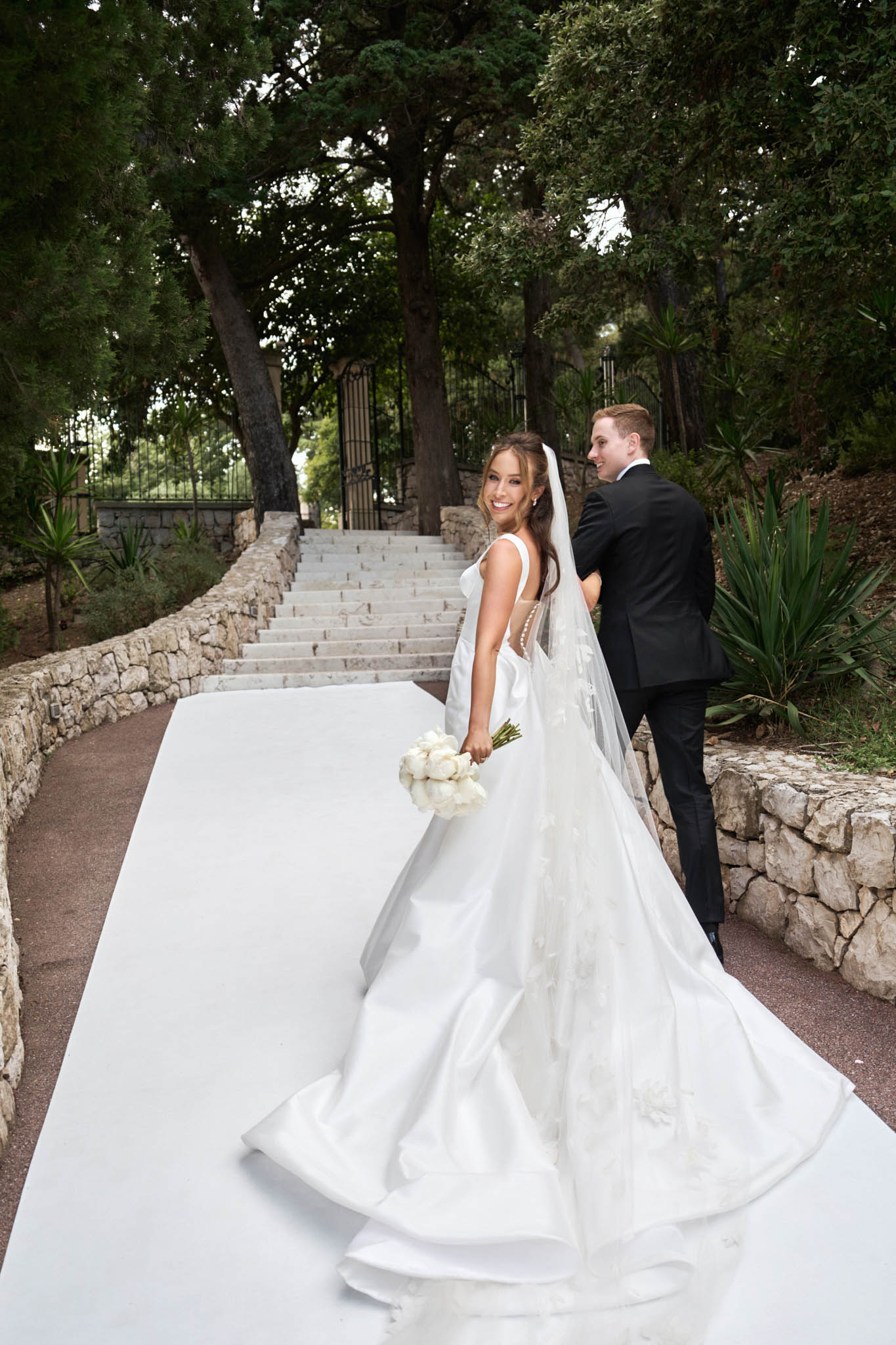 A couple walks along a white aisle runner toward a stone staircase at an outdoor venue, with the bride turning back to smile at the camera. The bride wears a white satin fit-and-flare gown with a deep back, pearl button detailing down the spine, and a very long cathedral veil with floral appliqué trim; she carries a round bouquet of white peonies. The groom is in a classic black suit and walks slightly ahead of her. The setting features rustic stone retaining walls lining the path, ornate wrought-iron gates at the top of the stairs, and dense mature trees overhead, consistent with a French villa or château garden. The shot is a medium-wide portrait taken from slightly behind and to the side, emphasizing the full length of the gown's train and veil against the white runner.