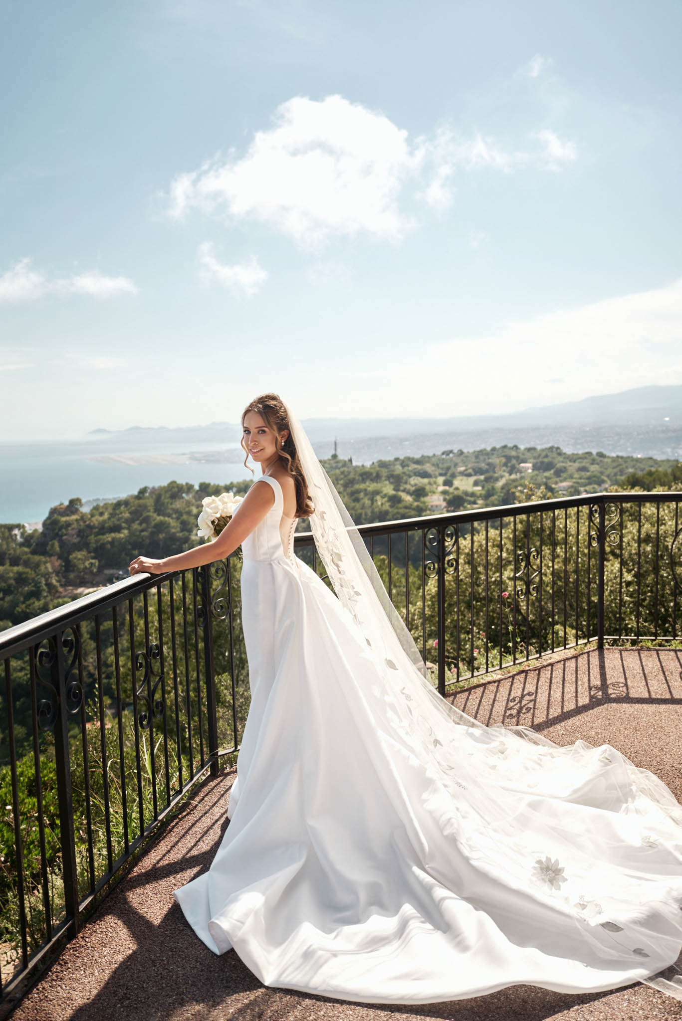 A bridal portrait taken outdoors on a terrace with wrought-iron scrollwork railings, overlooking a panoramic coastal landscape with a bay visible in the distance. The bride stands in three-quarter profile, glancing back toward the camera, wearing a white sleeveless A-line gown with a low back and an extended cathedral-length train that spreads across the terrace floor. She wears a long cathedral veil with scattered appliquéd floral details throughout its length. Her bouquet consists of white roses and she wears her dark hair in a half-up style. The overall styling is classic and clean, with a modern minimalist dress paired with an ornate veil. Full-length portrait shot in bright natural daylight.