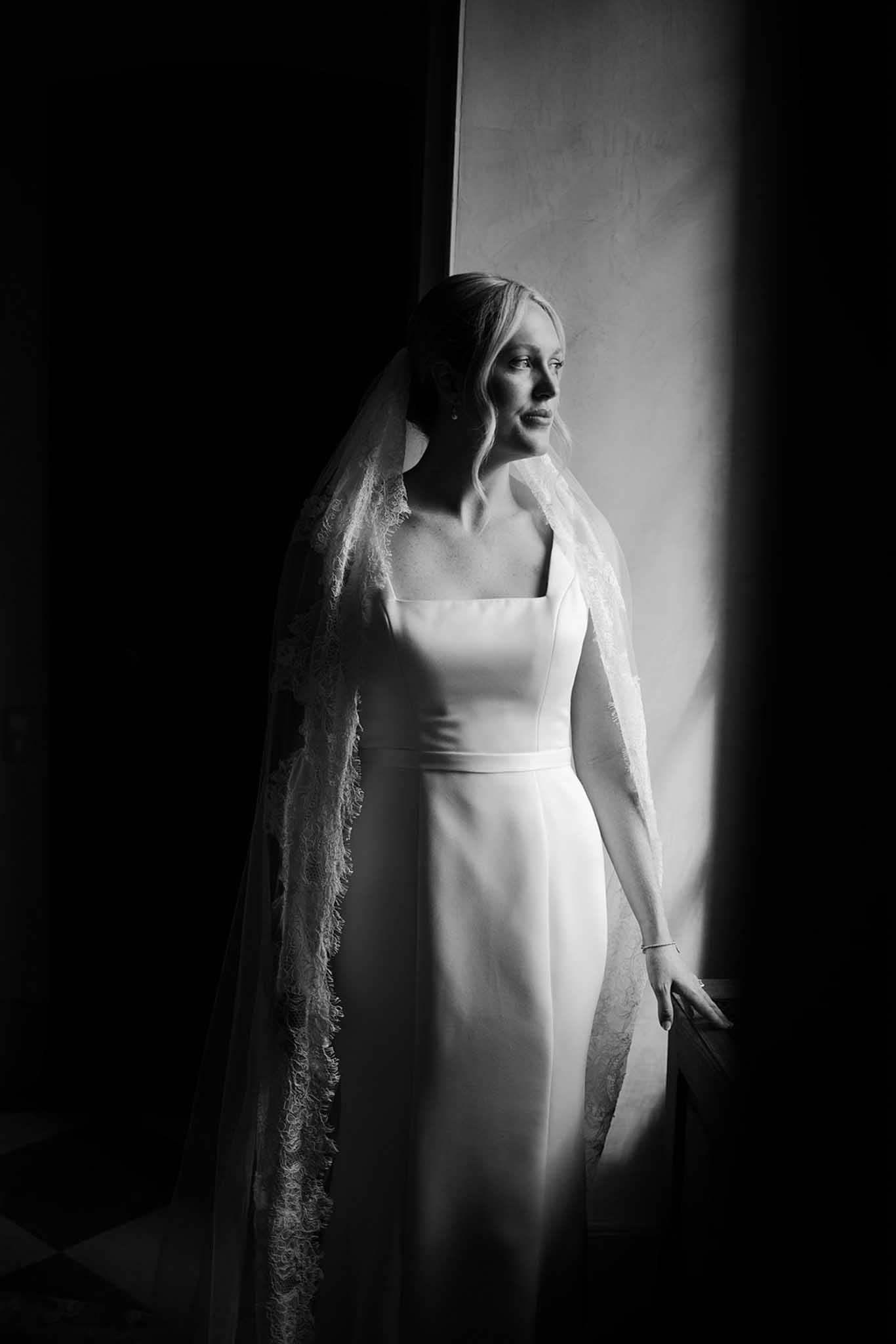 Black-and-white profile portrait of bride in minimalist dress and long lace veil with dramatic window light