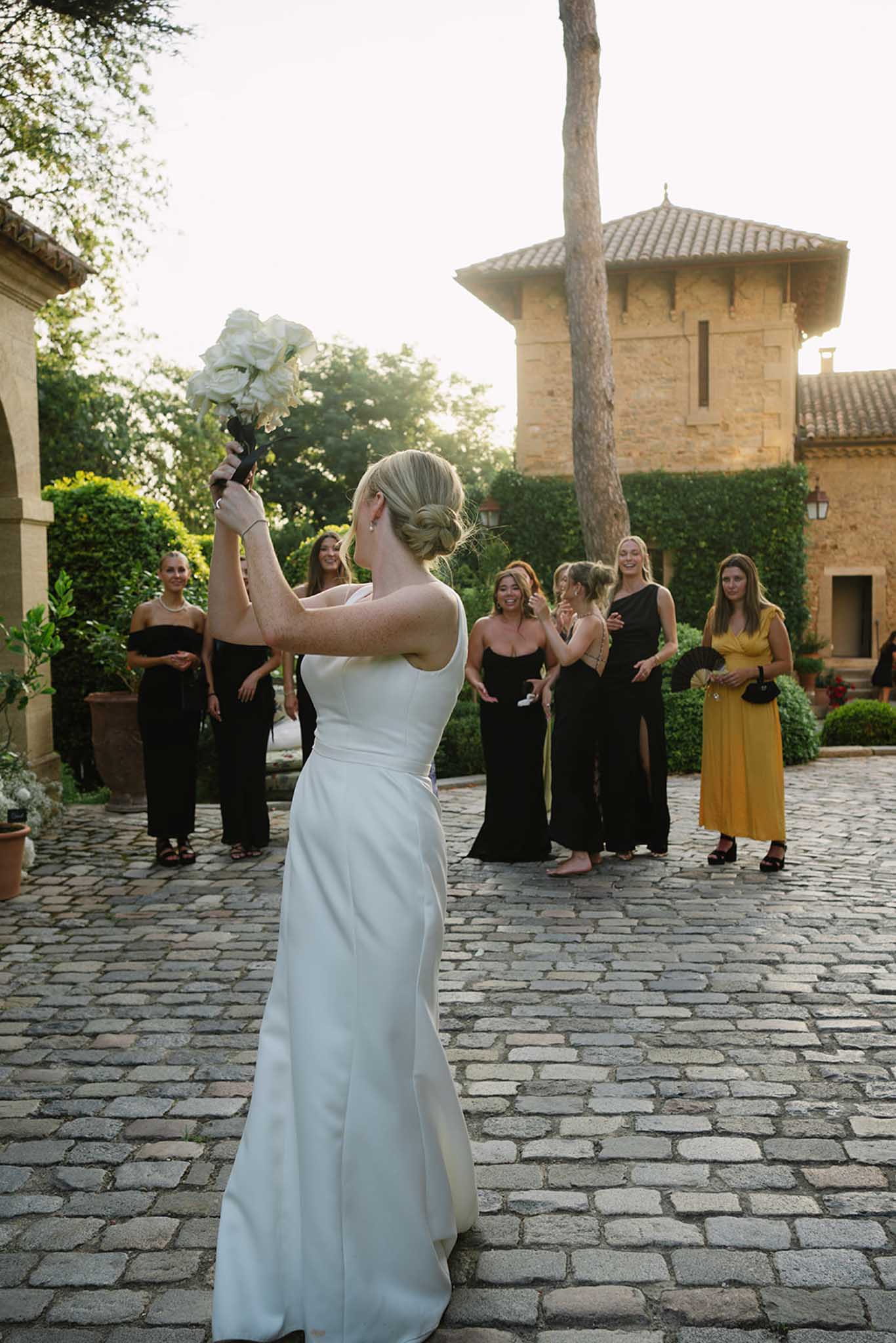 Bride in ivory gown performing bouquet toss at outdoor courtyard reception with guests in black gowns gathered around her