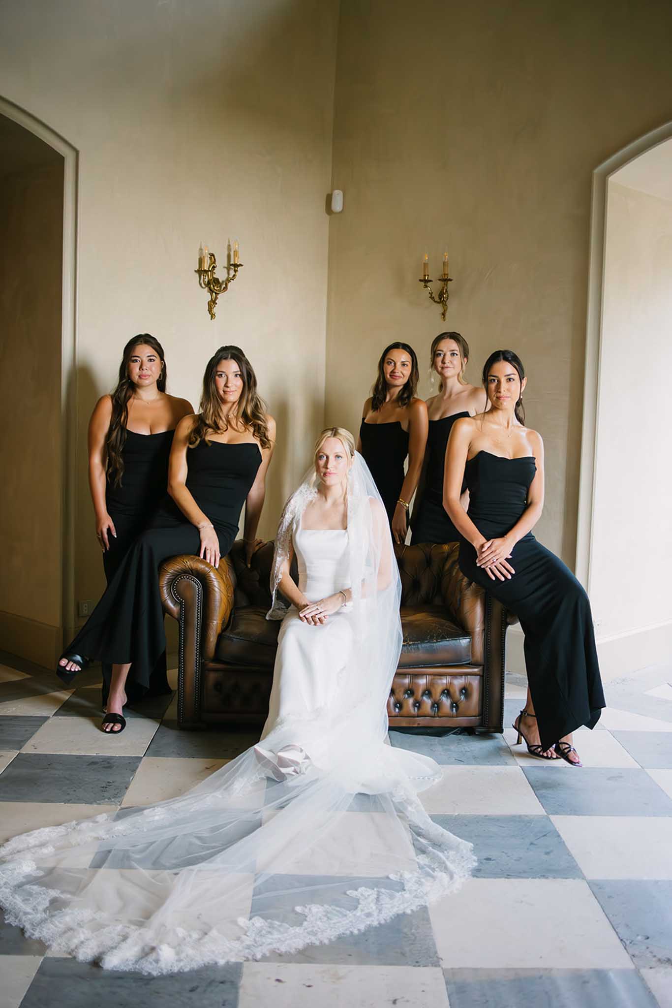 Bride in white gown and lace veil seated with five bridesmaids in black dresses on leather Chesterfield sofa