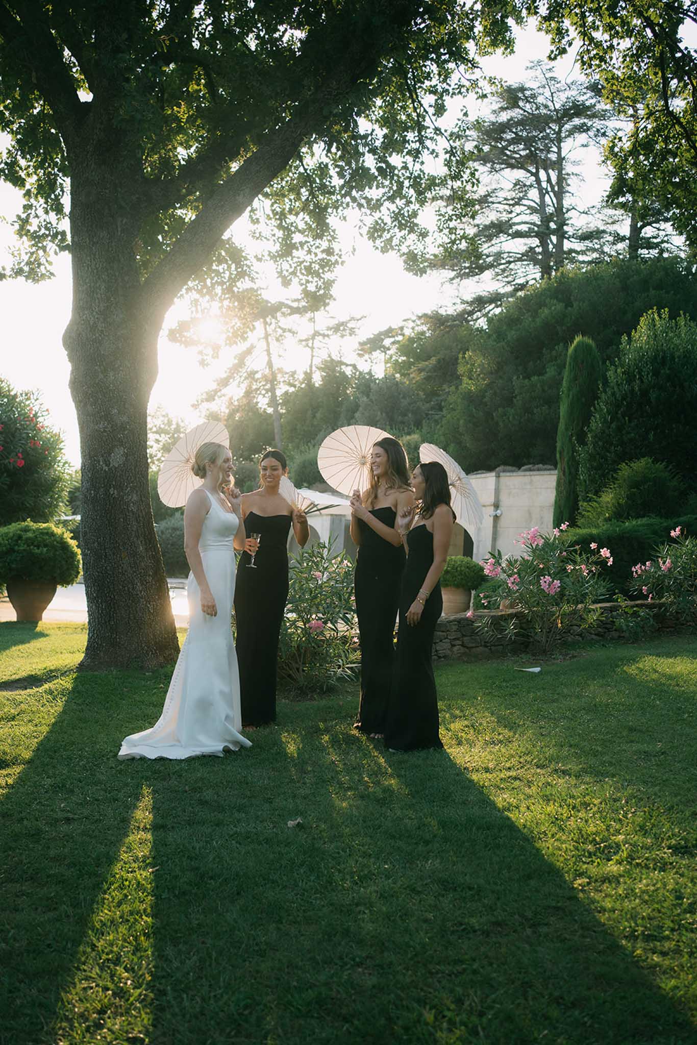 Bride and three bridesmaids in black gowns with white parasols beneath tree in garden