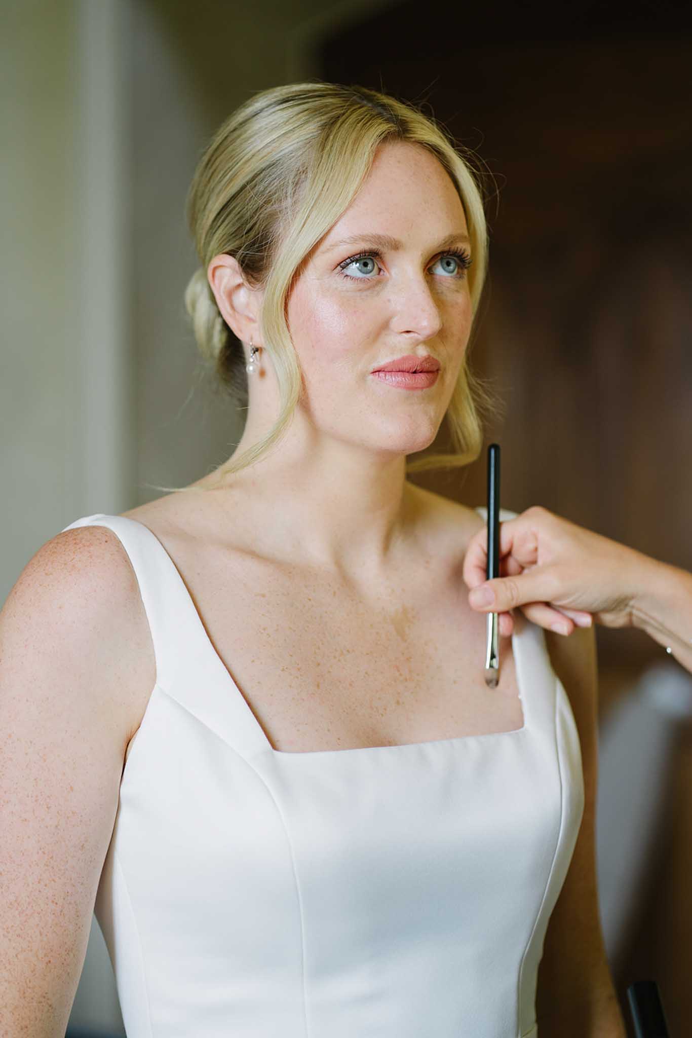 Bride in ivory dress holding a makeup brush during getting-ready preparations with soft background