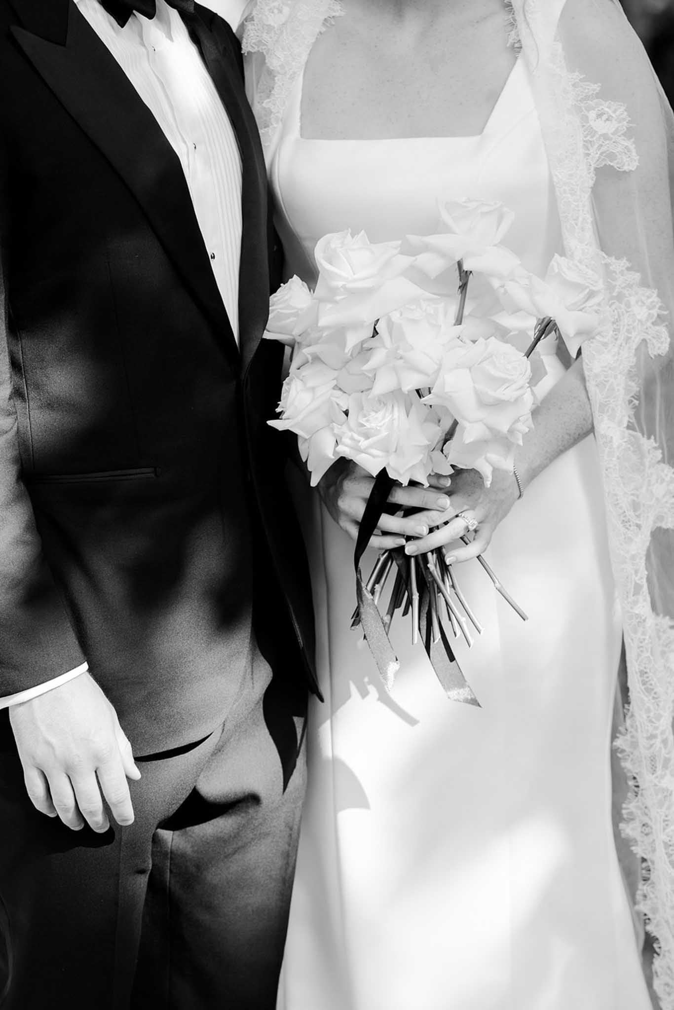 Black and white close-up of bride and groom showing bouquet of white roses, gloved hands, and formal attire