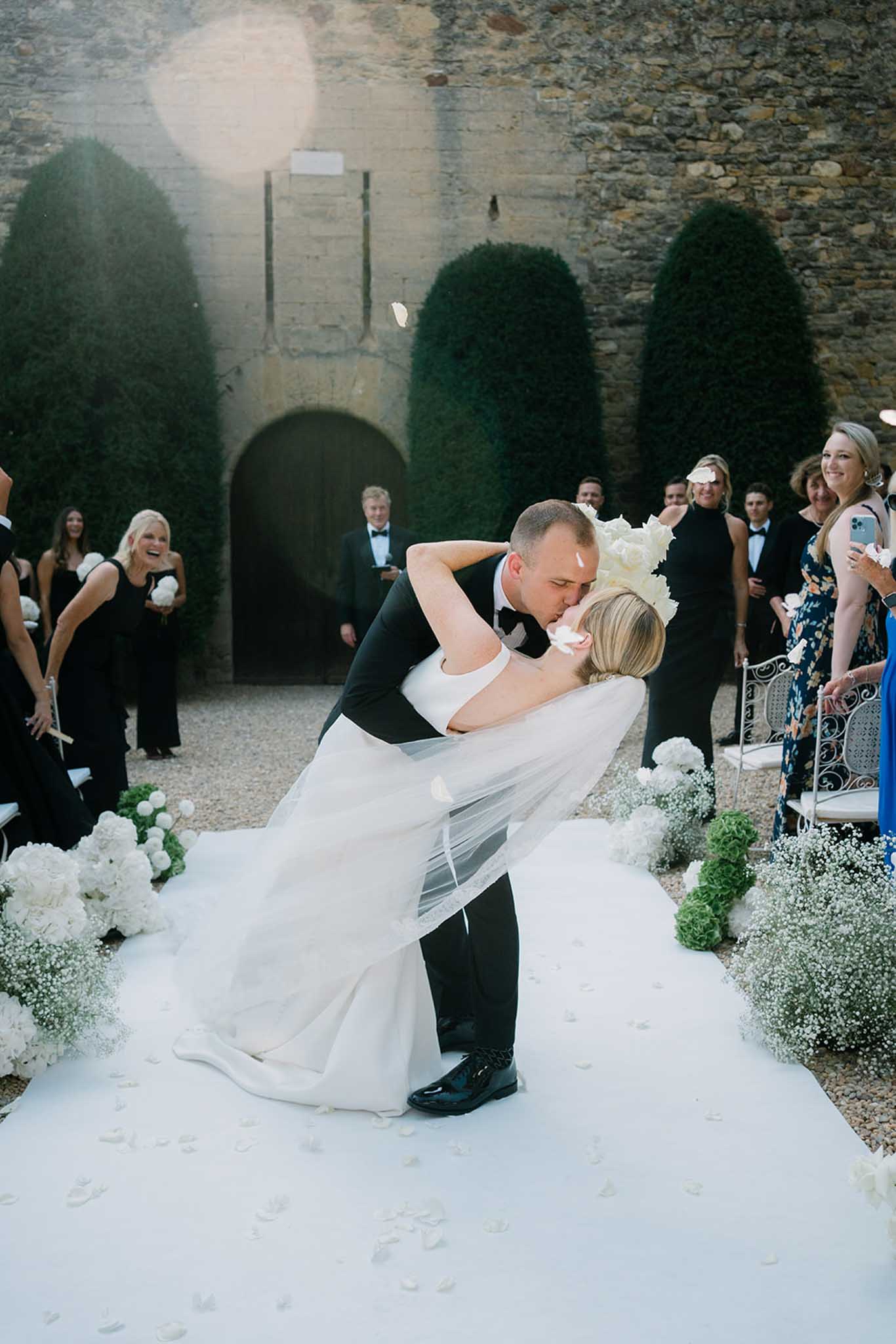 Groom dips bride during ceremony in stone courtyard with ivy-covered walls as guests watch in a semi-circle