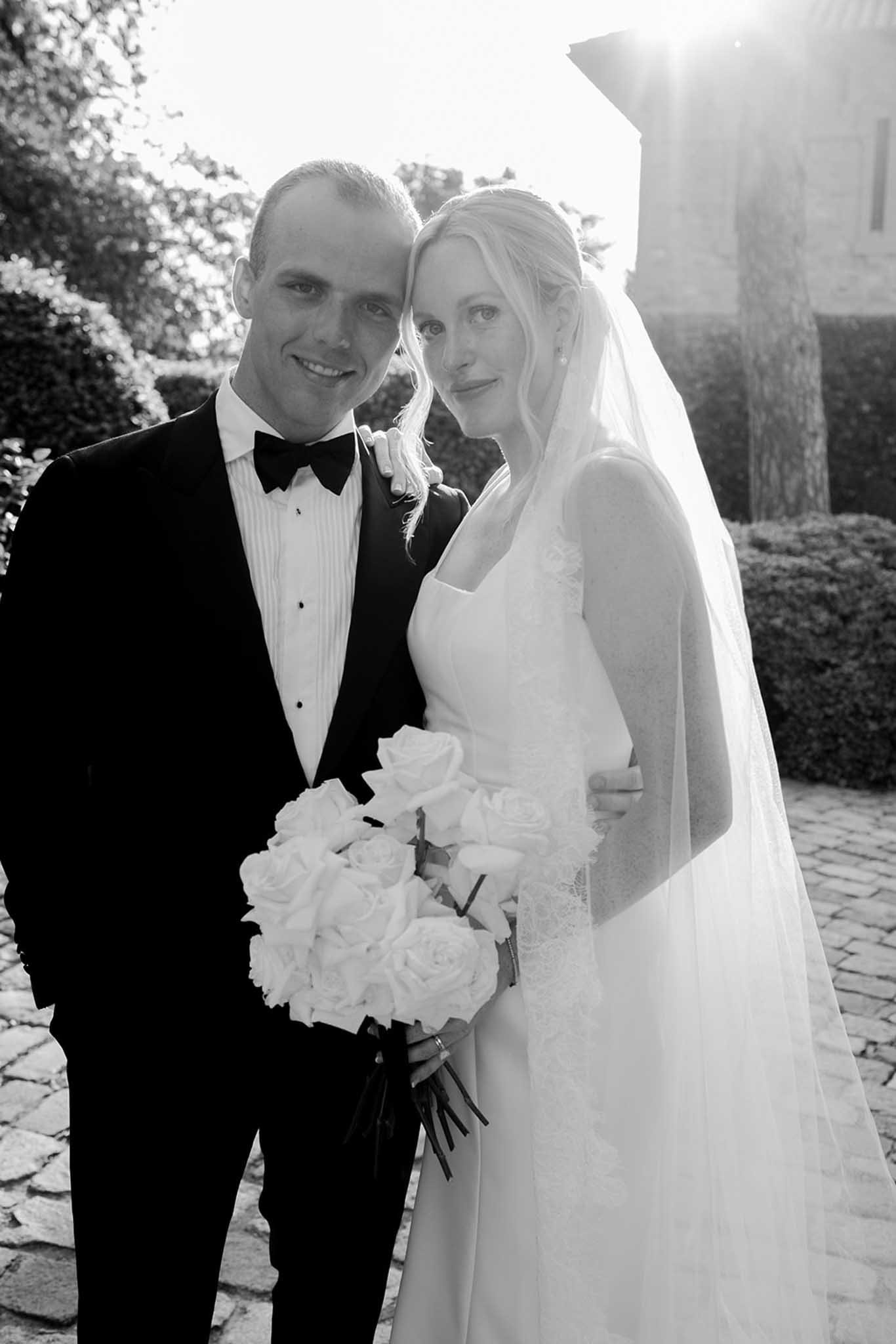 Bride in white gown and groom in black tie in formal garden, holding large white rose bouquet, black-and-white