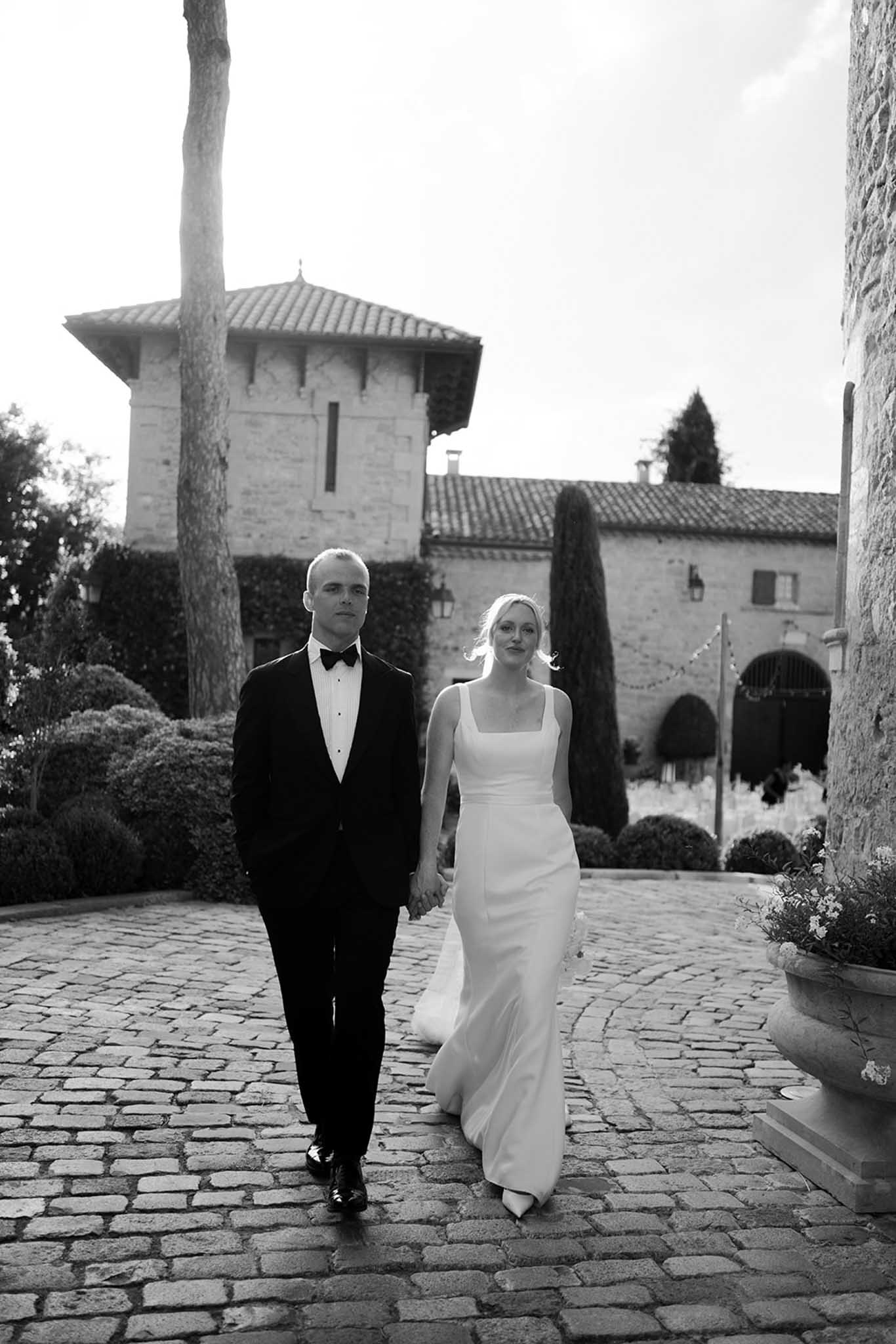 Black and white portrait of bride and groom walking through stone courtyard with tower behind them