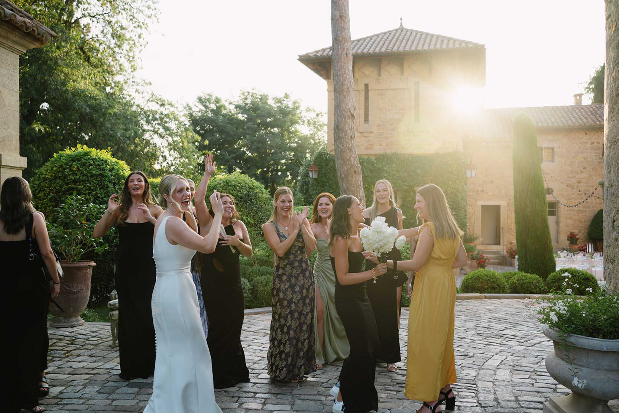 Bride tossing bouquet to twelve guests in stone courtyard with ivy-covered walls, cypress trees, and golden hour light