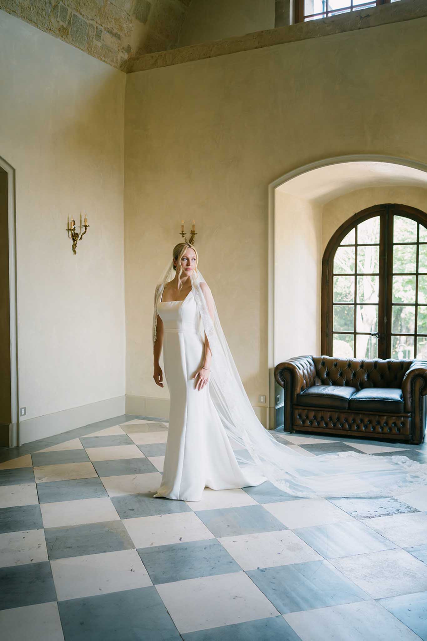 Bride in ivory column gown with floor-length veil standing in formal interior with arched windows and geometric tile floor