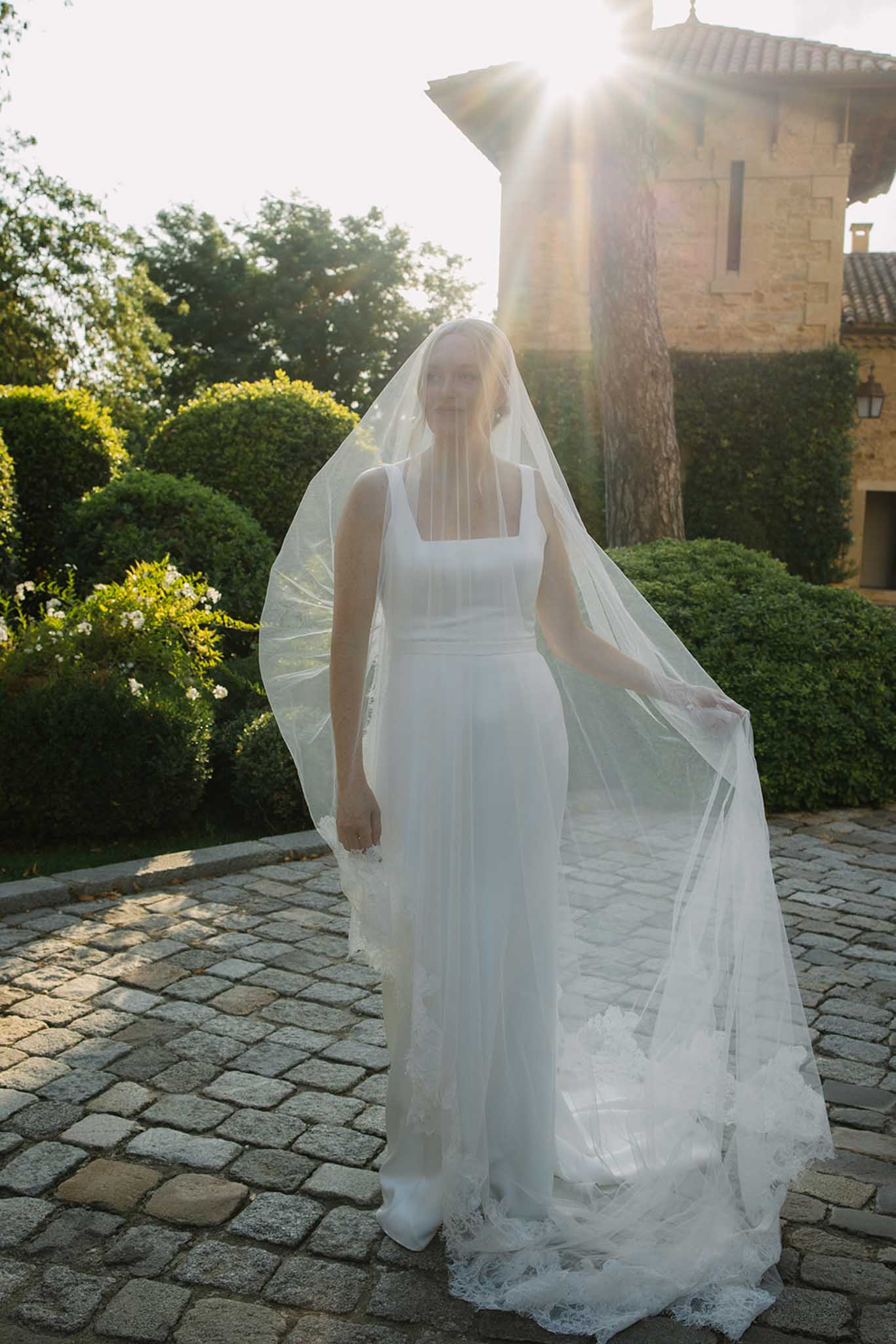 Bride in ivory column gown with long lace-hem veil stands in stone courtyard at golden hour with lens flare