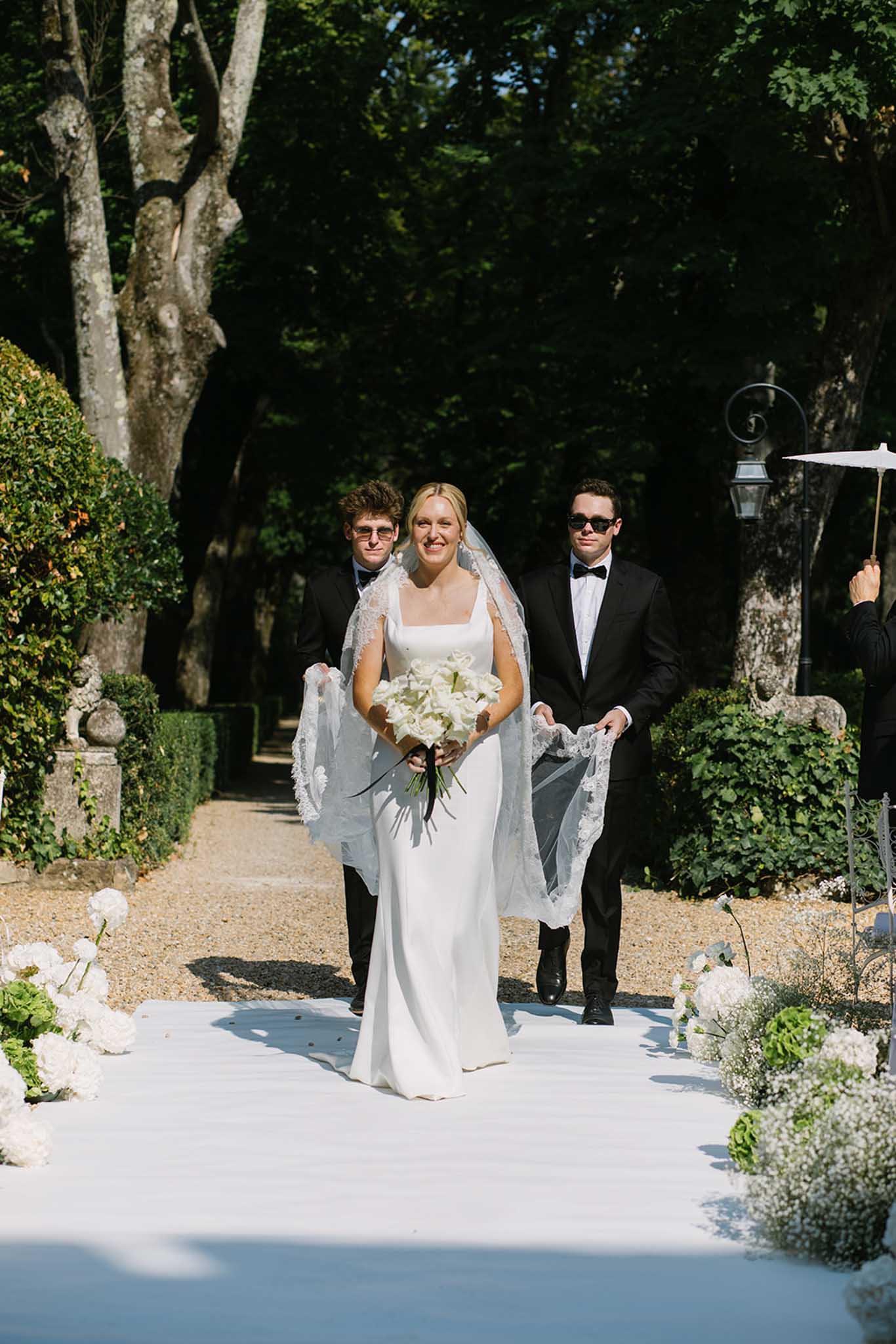 Bride walking down garden aisle with lace veil holding white hydrangea bouquet flanked by two groomsmen