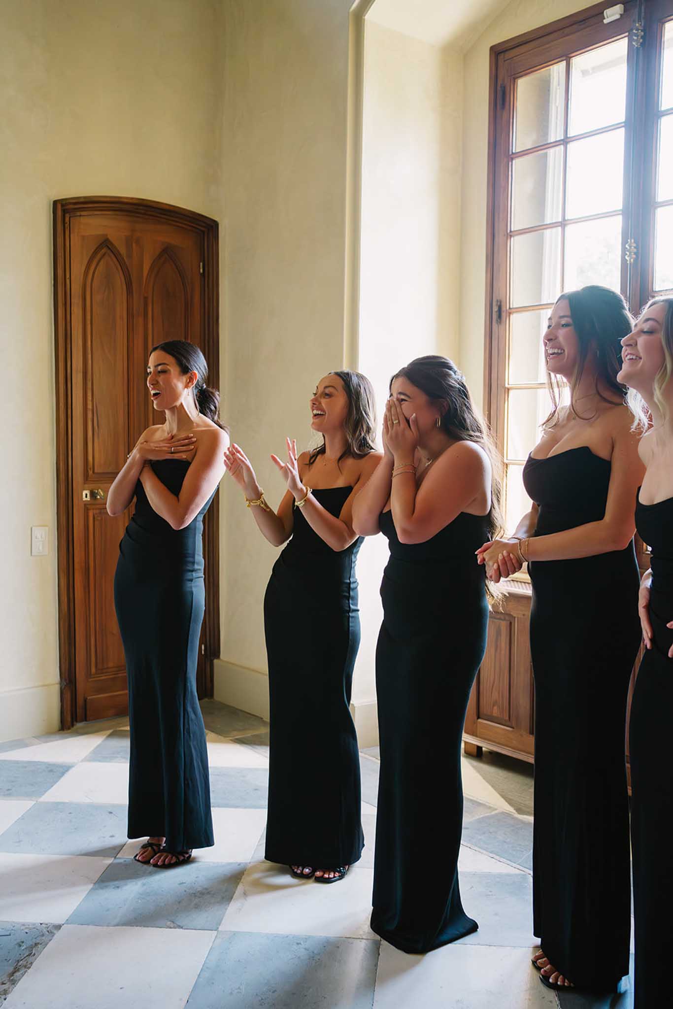 Five bridesmaids in black strapless gowns laughing together in a sunlit classical hallway with marble floor