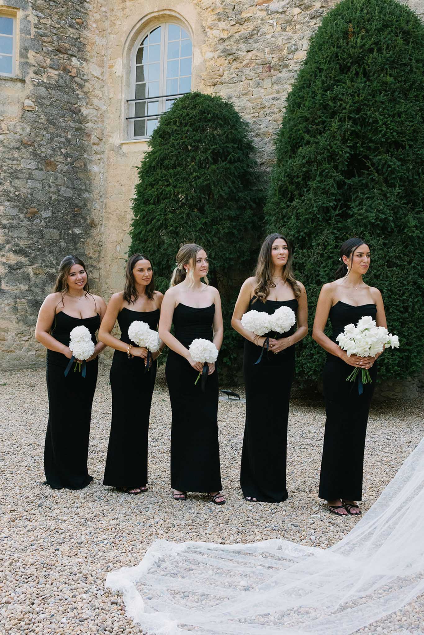 Five bridesmaids in matching black strapless dresses holding white hydrangea bouquets outside a stone building